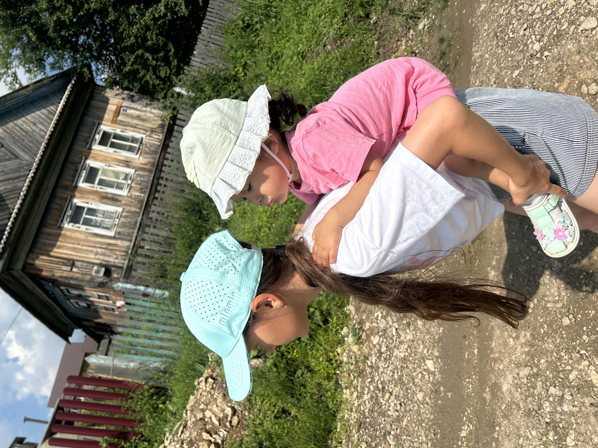 In Новомуллакаево, Russia, on a sunny day, a young girl with long dark hair, wearing a white t-shirt and striped shorts, carries a toddler on her back. The toddler, wearing a pink t-shirt and a light green hat with lace trim, looks to the side. The girl wears a light blue perforated baseball cap. They are walking on a dirt path next to a grassy area, with a weathered wooden house and a rustic fence in the background.