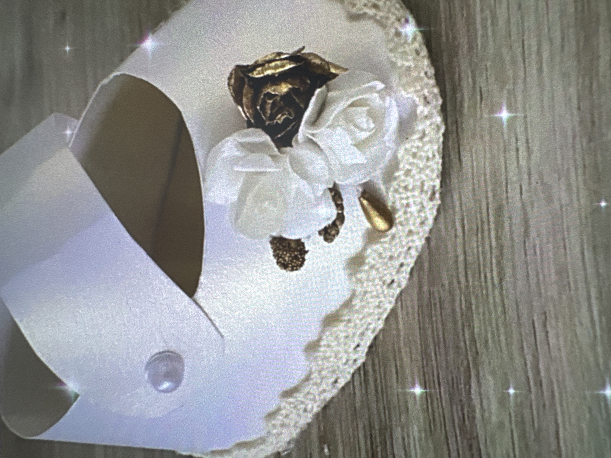 An overhead shot captures a single, ornate white baby slipper or sandal, positioned on a wooden surface that features prominent vertical grain. The slipper itself is crafted from a smooth, possibly satin-like white fabric, forming a broad strap across the top that is secured by a small, translucent, pearlescent button or bead.
The focal point of the slipper is a decorative cluster of three roses arranged on its upper surface. One rose, centrally placed, appears to be made of metallic material, possibly bronze or antique gold, with a slightly tarnished or textured finish. Flanking this metallic rose are two smaller, delicate white roses, fashioned from fabric, suggesting a soft and ethereal quality. Below these roses, a small, golden, droplet-shaped embellishment adds another touch of elegance.
The sole of the slipper is edged with a decorative trim, appearing to be white or cream-colored crochet or lace, featuring a scalloped pattern that adds to its delicate aesthetic. The wooden background, likely a floor or tabletop, shows variations in tone and texture. Numerous small, star-like light reflections, possibly lens flares or a digital effect, are scattered across both the slipper and the wooden surface, lending a whimsical or celebratory atmosphere to the image. No people or visible text are present, and no specific time of day or location cues are discernible beyond the wooden setting.