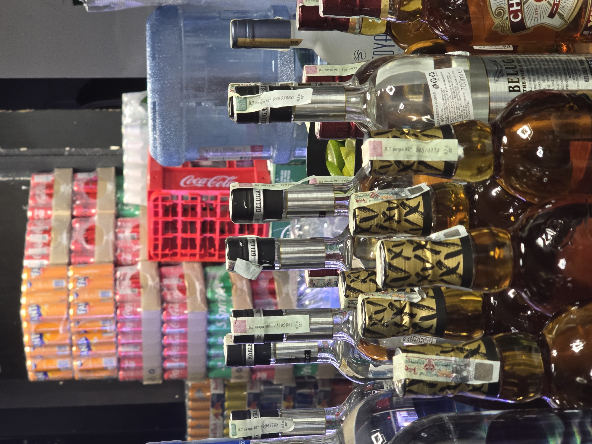 A stacked assortment of beverages is shown in this close-up photograph, likely taken in a retail environment in Plovdiv, Bulgaria. In the background, a red Coca-Cola crate is visible, as well as rows of Fanta and Sprite cans. In the foreground are rows of stacked liquor bottles, predominantly vodka, including brands like Beluga and other bottles with gold-colored details. Some bottles have excise tax stamps attached. A large water jug is situated behind the liquor bottles. The scene suggests a store shelf or storage area.