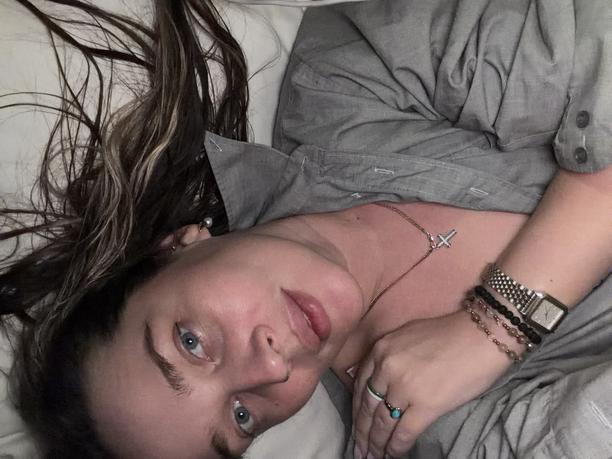 A woman with shoulder-length dark hair with lighter brown highlights lies on a white pillow in what appears to be a bedroom. She is wearing a grey button-down shirt. She has blue eyes and is looking directly at the camera. She has a nose ring and is wearing a pearl earring in her left ear. A necklace with a cross pendant is visible around her neck. On her right wrist, she is wearing an analog wristwatch with a metal band and two beaded bracelets. A ring with a turquoise stone is visible on her right hand. The image does not depict any elements suggesting it was taken in Moscow, Russia.