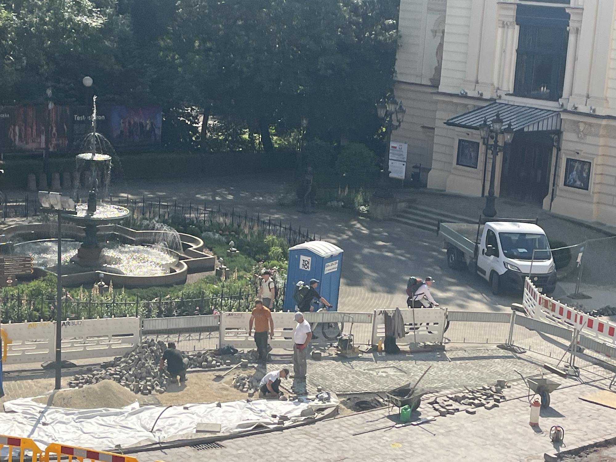The image shows an outdoor scene in a city, possibly undergoing construction or renovation. In the foreground, there is a large white tarp spread out on the ground, with piles of stones and sand around it. Several construction workers are visible, some of whom are wearing orange or white shirts. They appear to be engaged in laying down paving stones. There are wheelbarrows and construction tools scattered in the area.
Behind the construction site, a white portable toilet is positioned next to a fence. Further back, a fountain with multiple tiers is visible, with water spraying upwards. Beyond the fountain, there are trees and a building with a grand facade and entrance. A white truck is parked on the right side of the image, near the building's entrance. The overall impression is a busy urban area with ongoing public works.