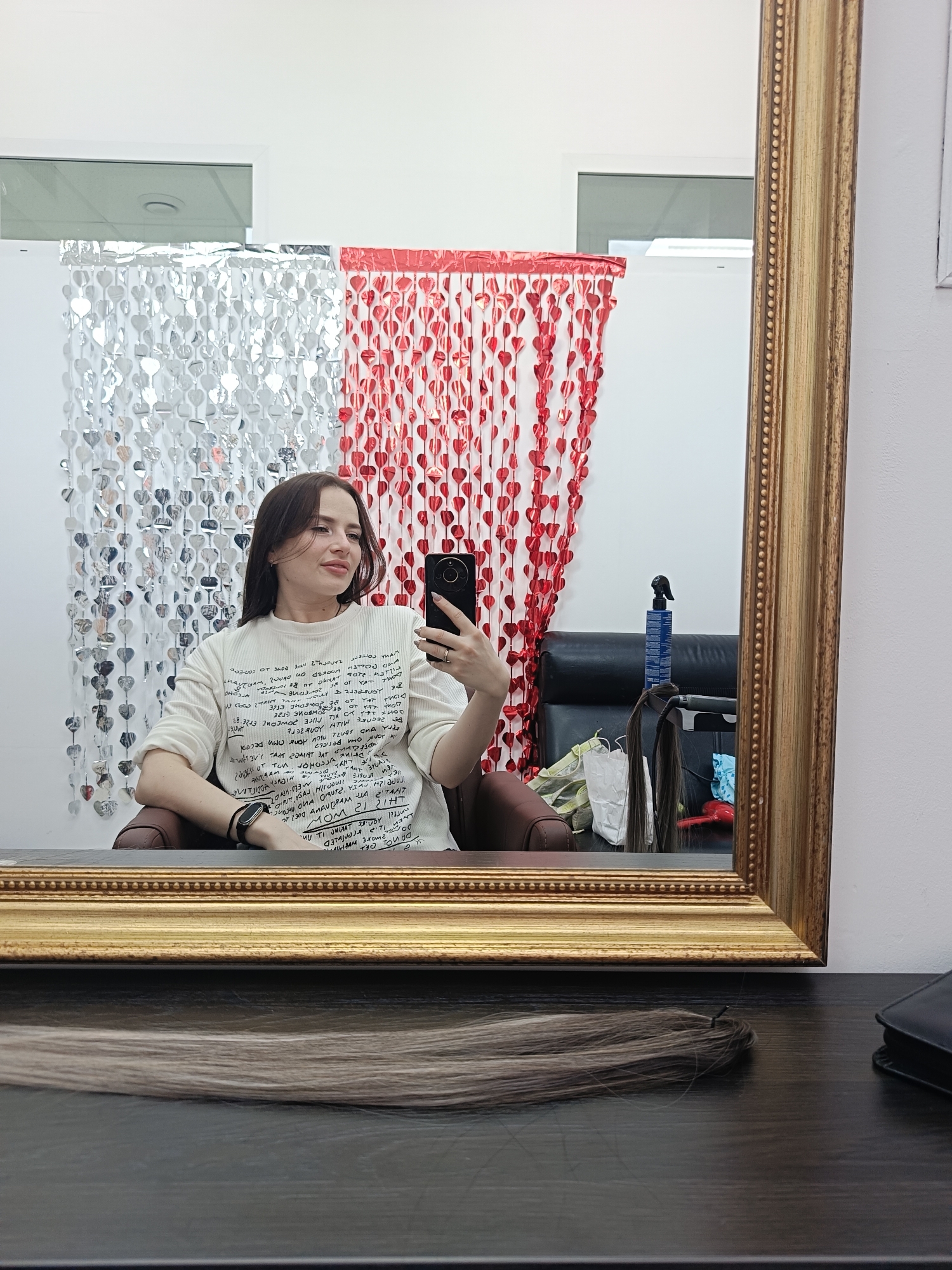 A young woman is captured in a mirror selfie within what appears to be a beauty salon, likely located in Chaikovsky, Russia. She is seated in a dark salon chair, holding a black smartphone to take the picture. She wears a light-colored, long-sleeved top adorned with stylized, mirrored text, much of which is illegible due to reflection and styling, though words like "MOM" and "THIS" are partially discernible, suggesting English content. A dark smartwatch is visible on her left wrist.
Behind her reflection, the wall is decorated with vertical strands of heart-shaped metallic decorations, silver on the left and red on the right, creating a festive backdrop. The mirror also reflects a black salon chair, a blue spray bottle, hair styling tools including a hair iron, and various white bags, indicating a workspace. Overhead, a window or opening shows a white ceiling.
On the dark wooden table in the foreground, a bundle of light brown hair extensions is laid out, along with a black wallet or clutch. The woman has a gentle, pleasant expression with a slight smile, looking to her right, away from the phone's lens. The overall setting is brightly lit, suggesting an indoor salon environment focused on hair care or styling, emphasized by the hair extensions on the table.