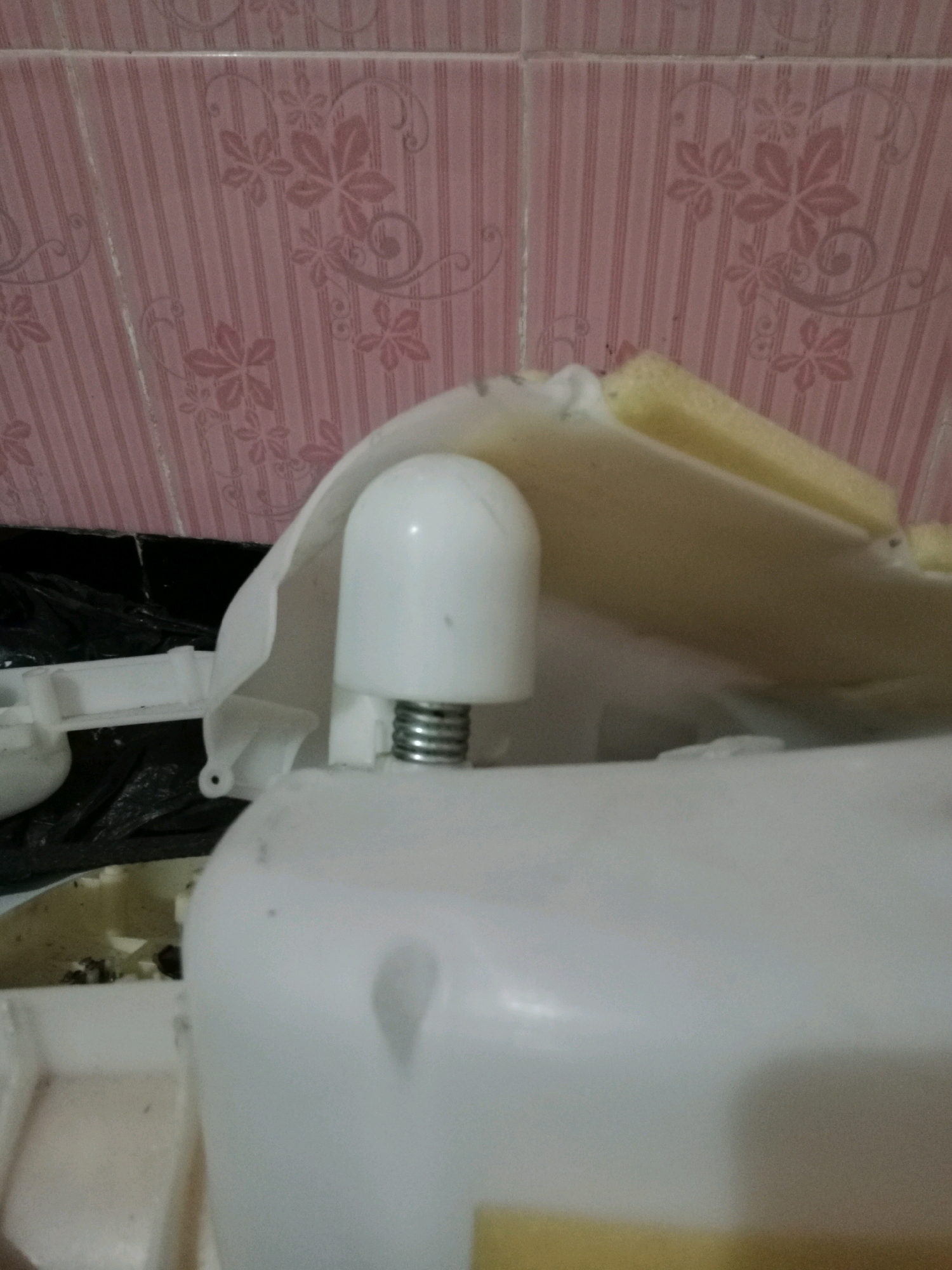 The image shows a close-up of a white plastic object with a silver spring protruding from its top. The object is positioned in front of a pink tiled wall with a floral pattern. To the left of the white object, a black plastic bag is visible. The overall scene appears to be an indoor environment, possibly a bathroom or a utility area, given the tiles. There is no information in the image to suggest a location in Ringinrejo, Indonesia.