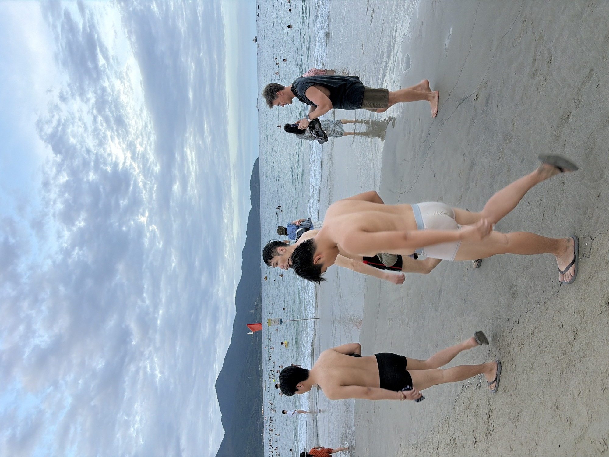 The image depicts a beach scene in Da Nang, Vietnam. In the foreground, three men are walking on the sand, two of them barefoot and one wearing black swim trunks and flip-flops. The third man from the left is wearing light-colored underwear and flip-flops. In the middle ground, a man in a black sleeveless shirt and khaki shorts is holding a phone and an object that appears to be a camera. Behind him, a woman in a patterned dress stands in the shallow water. The background shows a crowded beach with many people swimming and relaxing in the sea. Mountains are visible in the distance, under a cloudy sky.