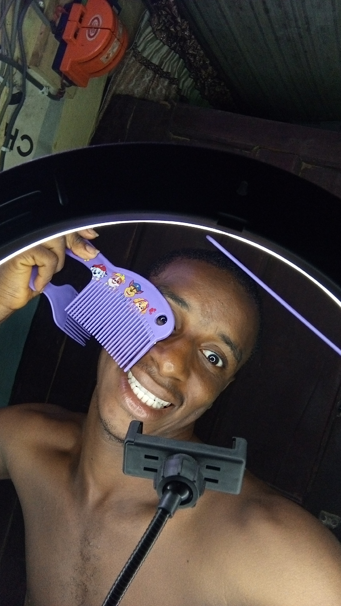 A young, dark-skinned man is captured in a close-up selfie or photograph, illuminated by a bright, circular LED ring light, part of which frames the top of his head. He smiles broadly, revealing white teeth, and looks directly at the camera with his left eye wide open, conveying a happy and playful mood. He is shirtless.
He holds a purple plastic comb, adorned with several Paw Patrol cartoon characters, playfully to the left side of his face. In the foreground, a black flexible arm mount with a smartphone clamp is visible, positioned below his chin, suggesting the device used for the photo.
The background depicts an informal, indoor setting, appearing somewhat dark and cluttered. To the upper left, an orange electrical cable reel or extension cord, along with various wires, are discernible. Below these, a light-colored surface shows the partial text "CH". Adjacent to this, a patterned fabric, possibly a curtain or bedding, with green, brown, and dark lace-like textures, is visible. On the right side of the background, a darker, textured surface is present. The overall scene suggests an indoor environment in Onitsha, Nigeria.