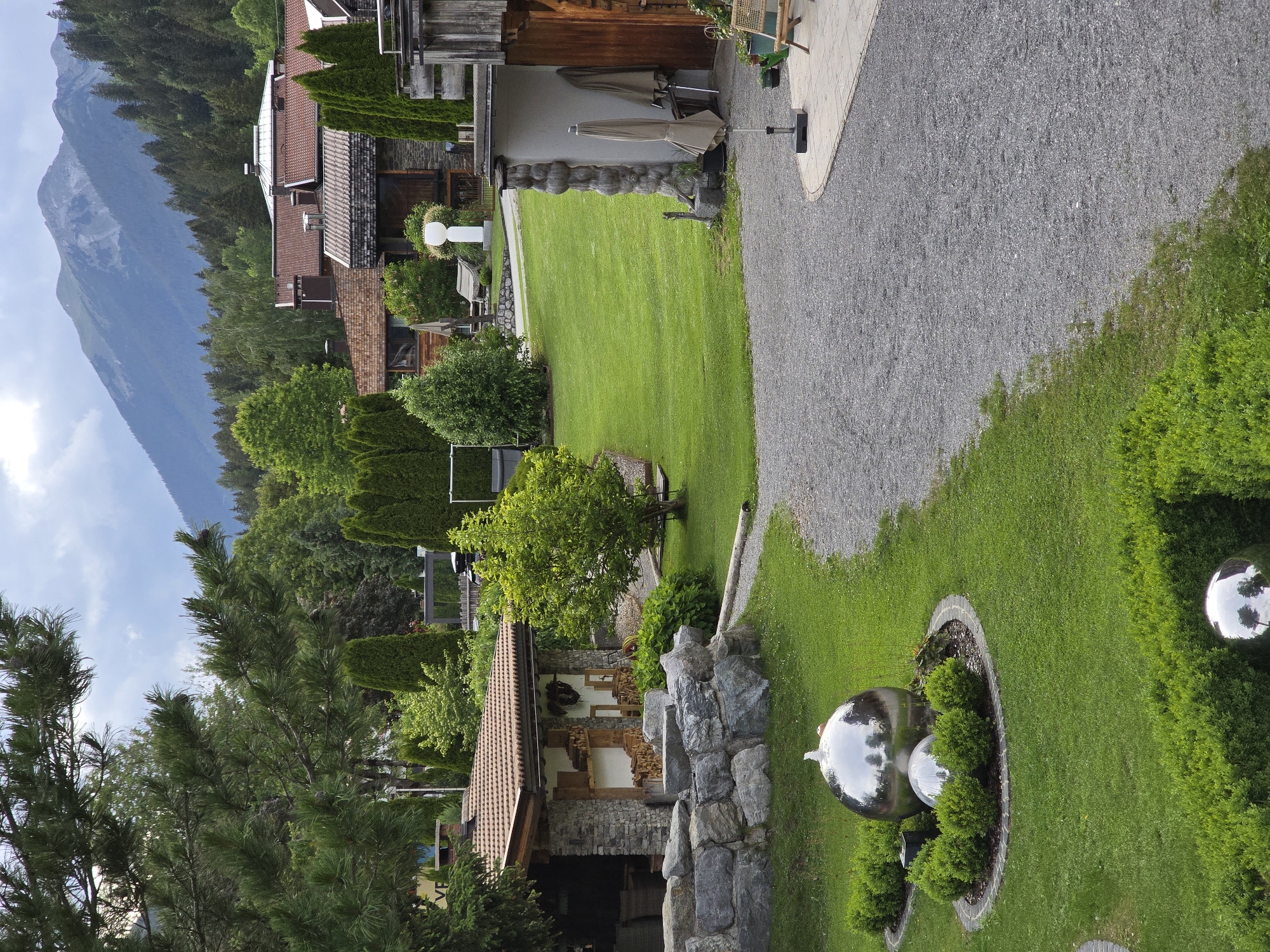 The image captures a serene, manicured outdoor space in Lermoos, Austria. In the foreground, a gravel path curves around a lush green lawn. Two prominent spherical metal sculptures are featured; the larger one, a water feature, is surrounded by neatly trimmed hedges and smaller spherical sculptures. Further into the scene, a rustic stone structure with a tiled roof displays firewood stacked neatly. Adjacent to it, a well-maintained lawn stretches towards a collection of buildings with terracotta tiled roofs, nestled against a backdrop of dense green trees and a majestic mountain range under a cloudy sky. Umbrellas, outdoor furniture, and potted plants suggest a recreational or hospitality setting. The overall impression is one of tranquility and natural beauty, characteristic of the Austrian Alps.