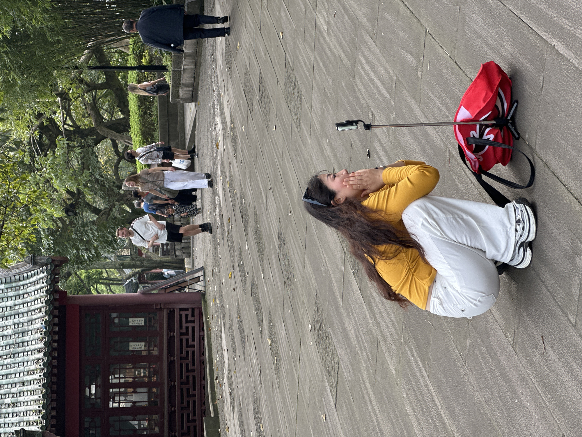 A wide, angled shot depicts a scene in Yuzhong District, China. A woman with long, dark hair, wearing a mustard yellow top and white pants, is crouched on a stone-paved area, her hands near her face as if posing for a picture. Next to her is a red umbrella and a selfie stick with a smartphone attached. In the background, a group of people walk along the same stone path. A traditional Chinese building with a tiled roof and intricate wooden details is visible on the left. Above the building and the stone walkway are tall, green trees that provide partial coverage for the people. The sky appears overcast. A man is visible near the top of the image near the center, as well as a short stone railing.