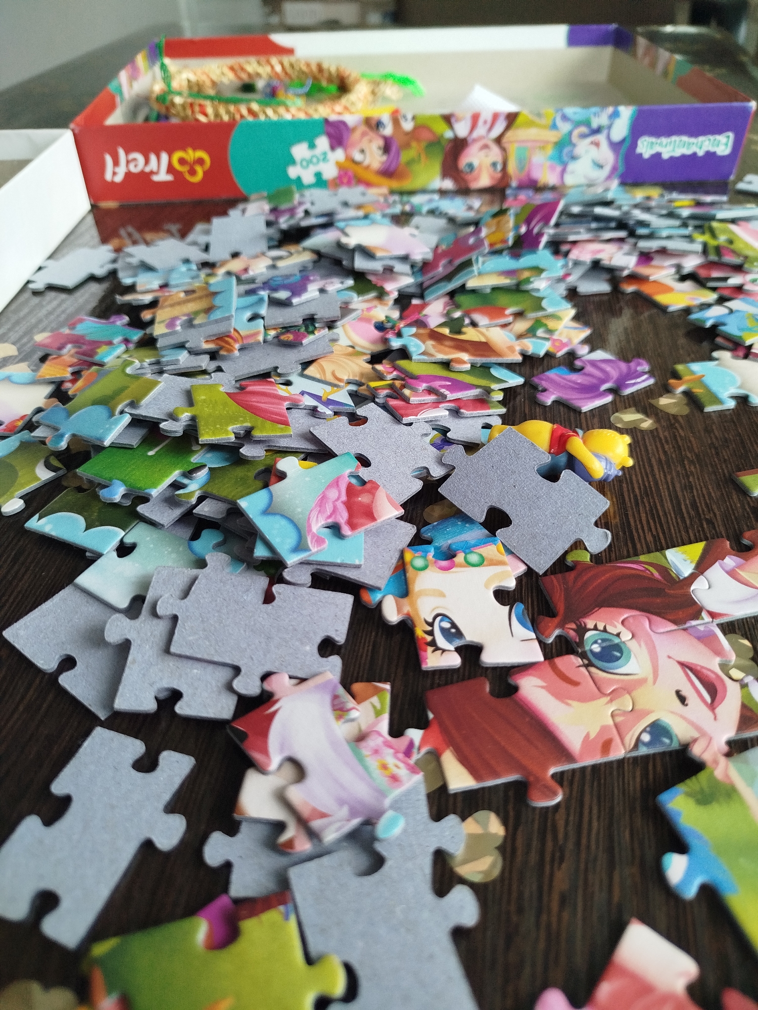 The image depicts a pile of scattered puzzle pieces on a wooden surface. The puzzle appears to feature characters from "Enchantimals" and possibly "Winnie the Pooh". Some of the pieces are gray, indicating they are border or background pieces, while others are brightly colored and feature parts of the characters' faces and clothing. The puzzle box, with the "Trefl" logo and "200" pieces indicated, is visible in the background. The scene is domestic, suggesting a home environment where a puzzle is being assembled. There are no people visible in the image, and the focus is solely on the puzzle pieces and the box. The wooden surface has a visible grain, indicating a tabletop or floor. The lighting suggests an indoor setting, likely with ambient light. The location of Крывасельскі сельскі Савет, Belarus is not depicted or implied in this image.