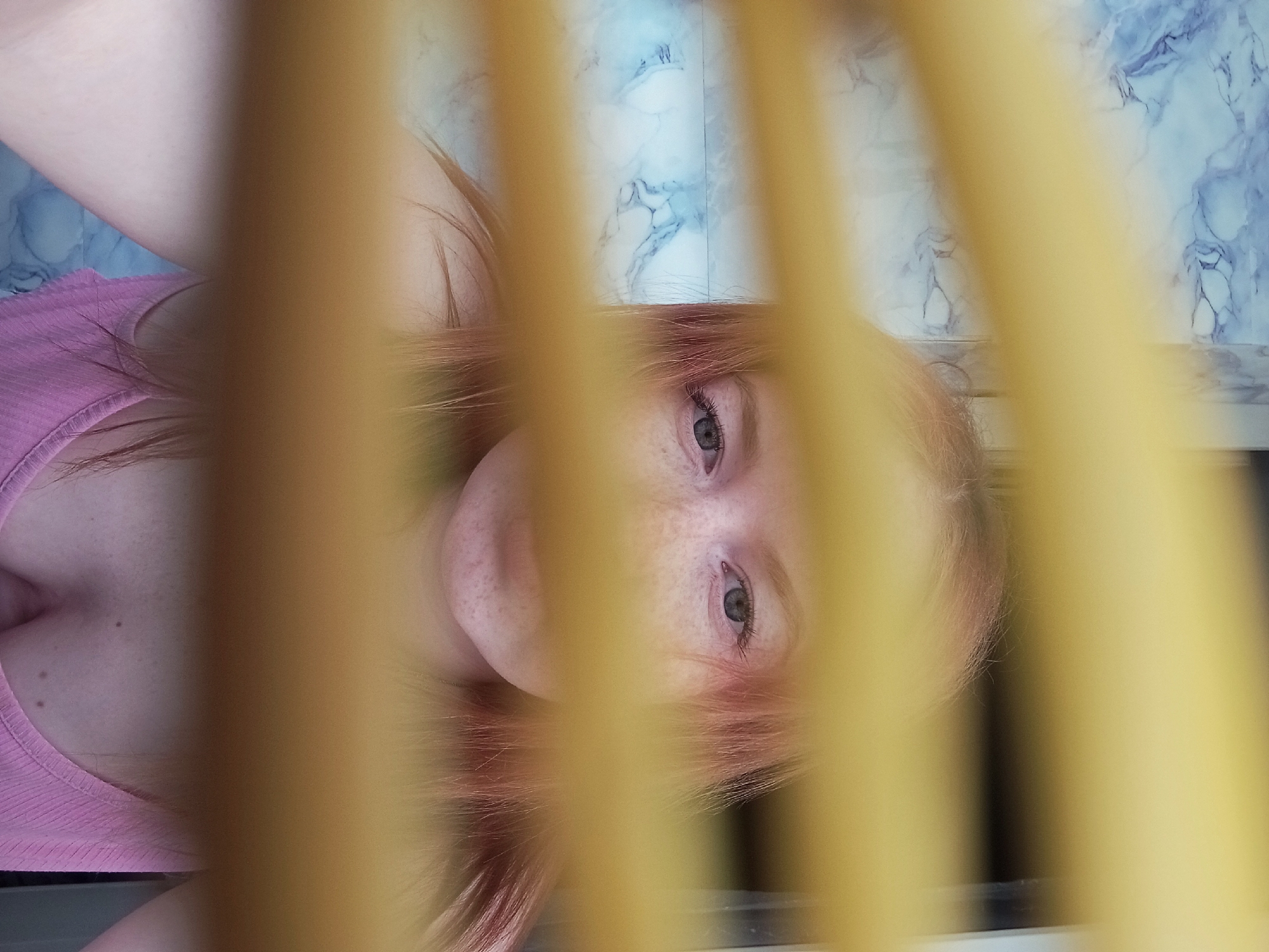 A fair-skinned individual with freckles and reddish-blonde hair is pictured from a low angle, partially obscured by vertical yellow bars, possibly a railing. The person wears a pink ribbed tank top. A patterned surface, possibly a wall or floor with blue and white marbling, is visible in the background. The bottom of a dark-colored structure, maybe a windowsill, is visible at the bottom of the image. The location is not discernible from the image.
