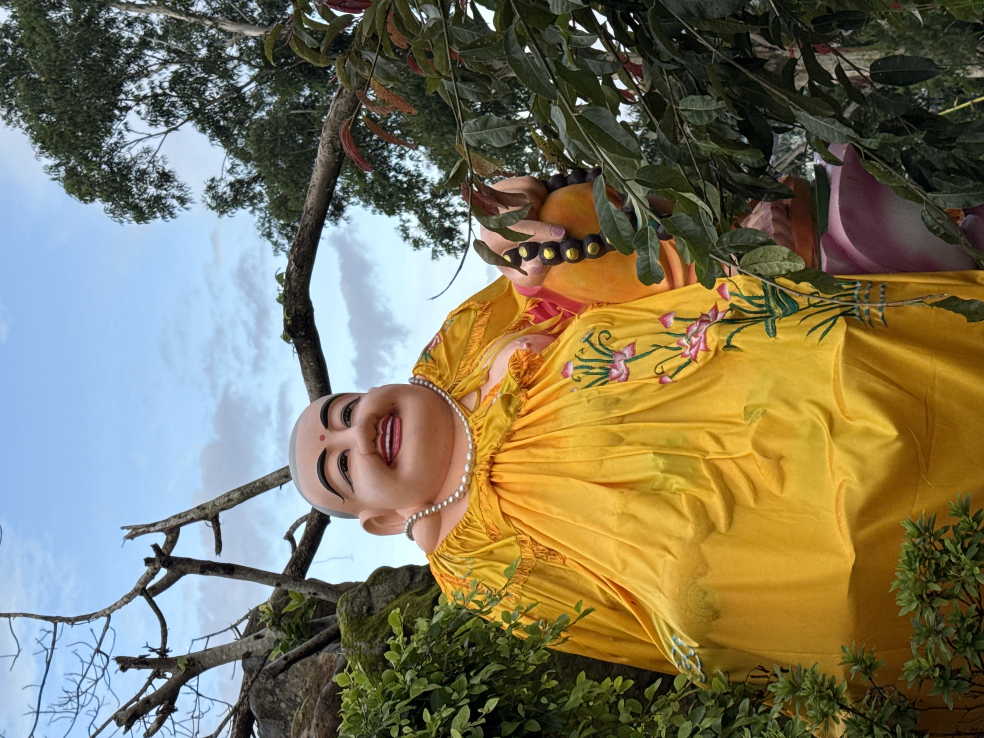 A large, colorful statue of a smiling Buddha is situated outdoors amidst lush greenery and trees under a cloudy sky. The Buddha is adorned in a bright yellow robe with embroidered pink flowers and wears a pearl necklace. Its right hand is raised and holding a string of dark beads. The statue is partially obscured by branches and foliage from surrounding trees and bushes. The cloudy sky suggests it is daytime. The location is identified as Lâm Viên - Đà Lạt, Vietnam.