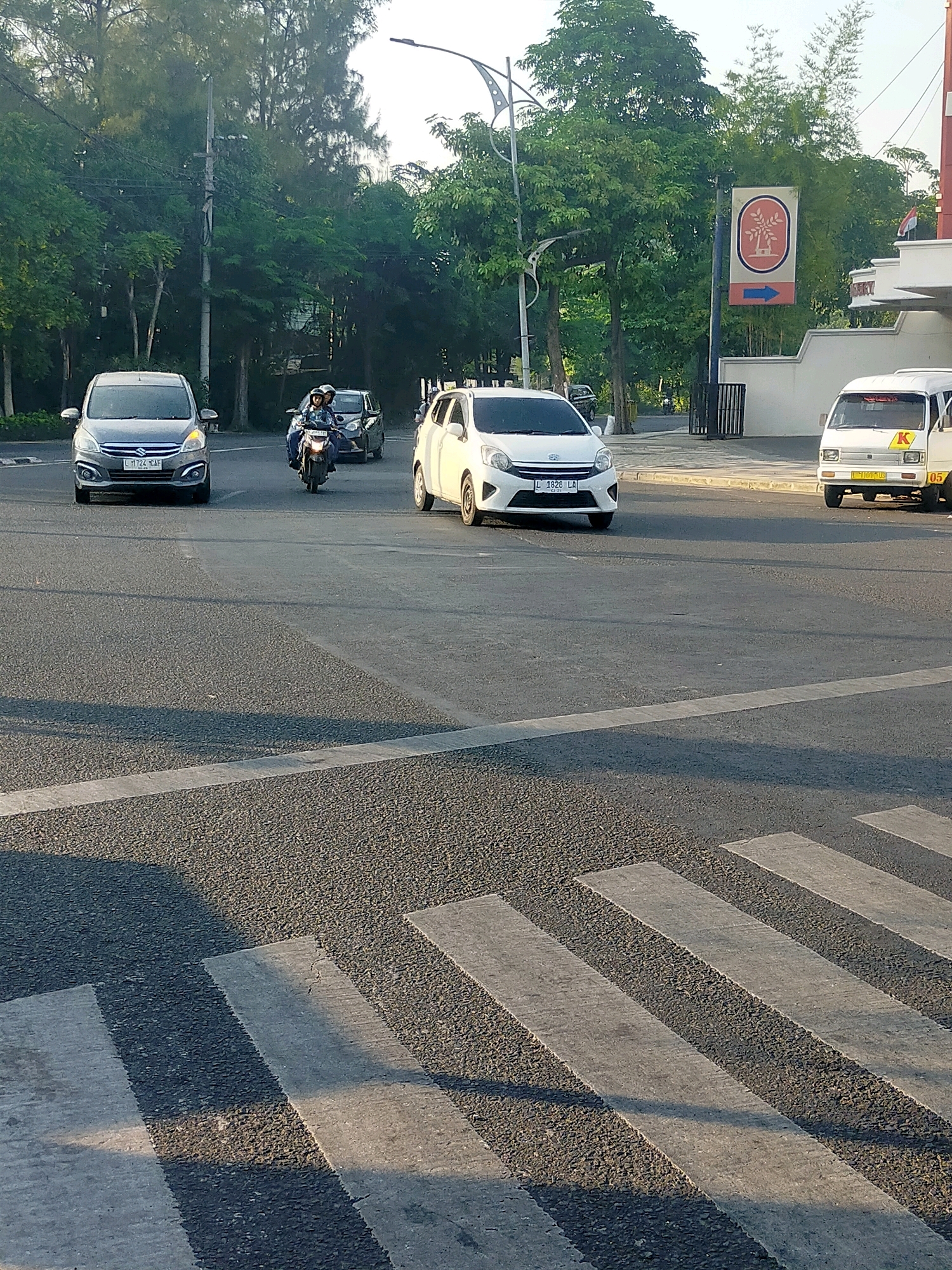 The image depicts a street scene in Surabaya, Indonesia, likely at an intersection with a pedestrian crossing. In the foreground, a zebra crossing is visible on the asphalt road. The middle ground shows several vehicles on the road. A gray Suzuki Ertiga car is on the left, its headlights on, and a license plate reading "L 1724 XAF". A motorcycle with two occupants, a driver and a passenger, is in the center, moving forward. To the right of the motorcycle is a white car, possibly a Toyota Agya, with a license plate "L 1828 LR". Further to the right, a small white van with a yellow "K" and the number "05" on its side is parked or moving slowly. In the background, lush green trees line the street, and a sign with a circular logo featuring a stylized tree and an arrow pointing left is mounted on a pole. A building with white walls and a modern architectural style is also visible. The sky is bright, indicating daytime, and the sun casts shadows on the road. The overall environment suggests a typical urban setting in Surabaya during daylight hours.