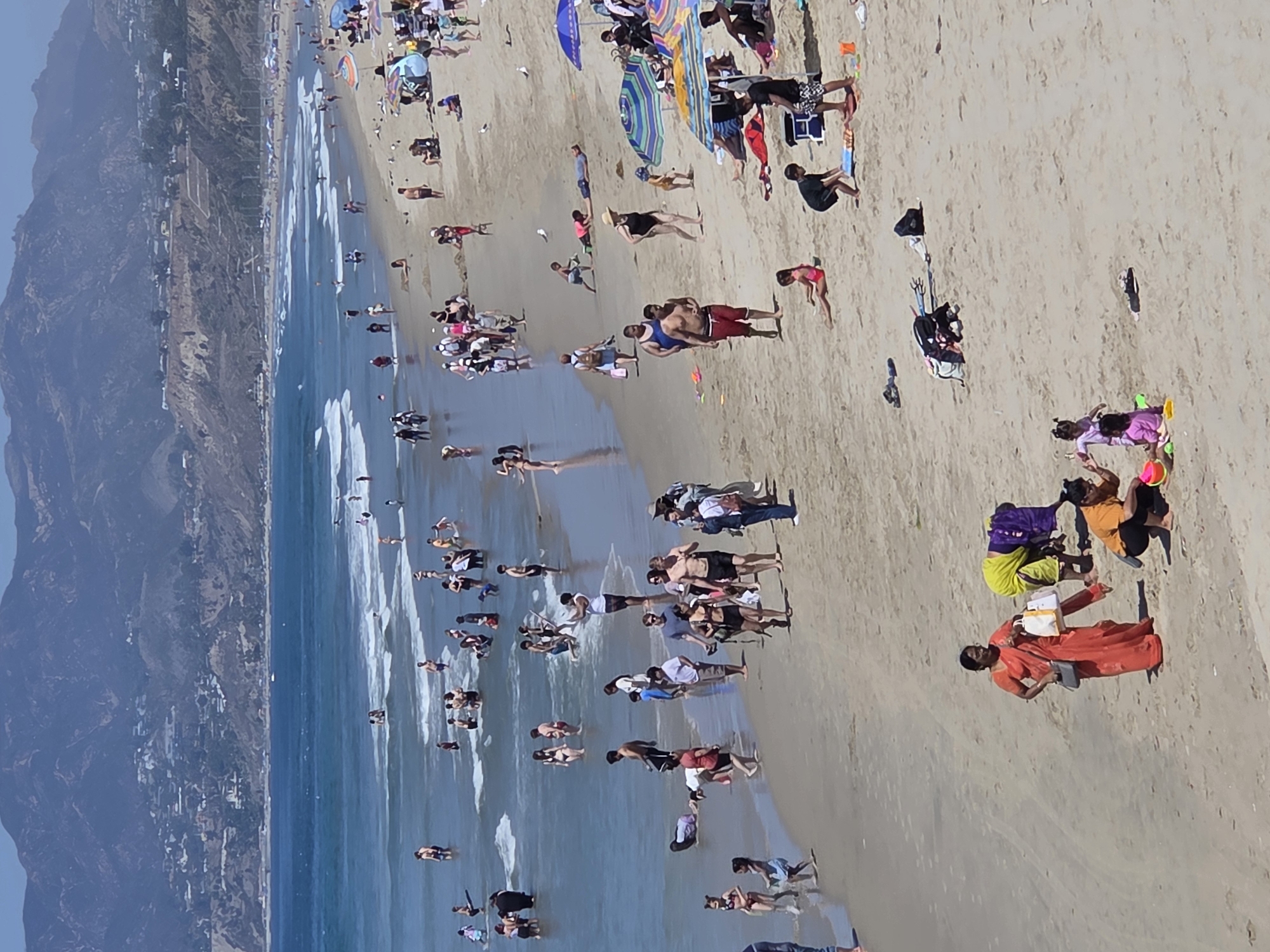 This is an outdoor, high-angle, full-shot photograph of a crowded beach in Santa Monica, California. The scene is dominated by a wide expanse of sandy beach meeting a blue ocean with gentle waves. In the background, a hazy, mountainous landscape rises behind the city, which appears as a collection of buildings and roads on the hillsides. The sky is clear and blue.
The beach is populated with numerous individuals engaged in various activities. In the water, people are wading, swimming, and standing. Closer to the shore, many are walking along the wet sand or playing. Further up the beach, groups are sunbathing, sitting on towels, and interacting. Umbrellas are dotted across the sand, providing shade. There are also several beach bags and other personal items visible.
Specific individuals and groups can be observed. Near the bottom of the frame, a woman in an orange sari stands, holding bags. Nearby, another woman in a yellow and purple garment bends over. Two young children are playing in the sand with toys. Further up the beach, a man in red shorts walks towards the water, while a woman in a black swimsuit walks away. Several other groups are scattered throughout the scene, with individuals standing, walking, and sitting. The overall impression is one of a lively and popular summer day at the beach.