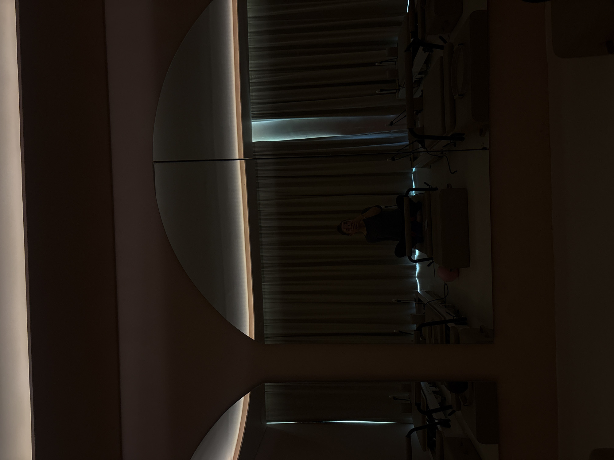 A woman's reflection appears in a segmented mirror that appears to be mounted on a wall. The mirror provides a view into a room with vertical blinds. The wall adjacent to the mirror is light pink, while the perpendicular wall is dark brown. The location is presumed to be in Mexico City, Mexico.