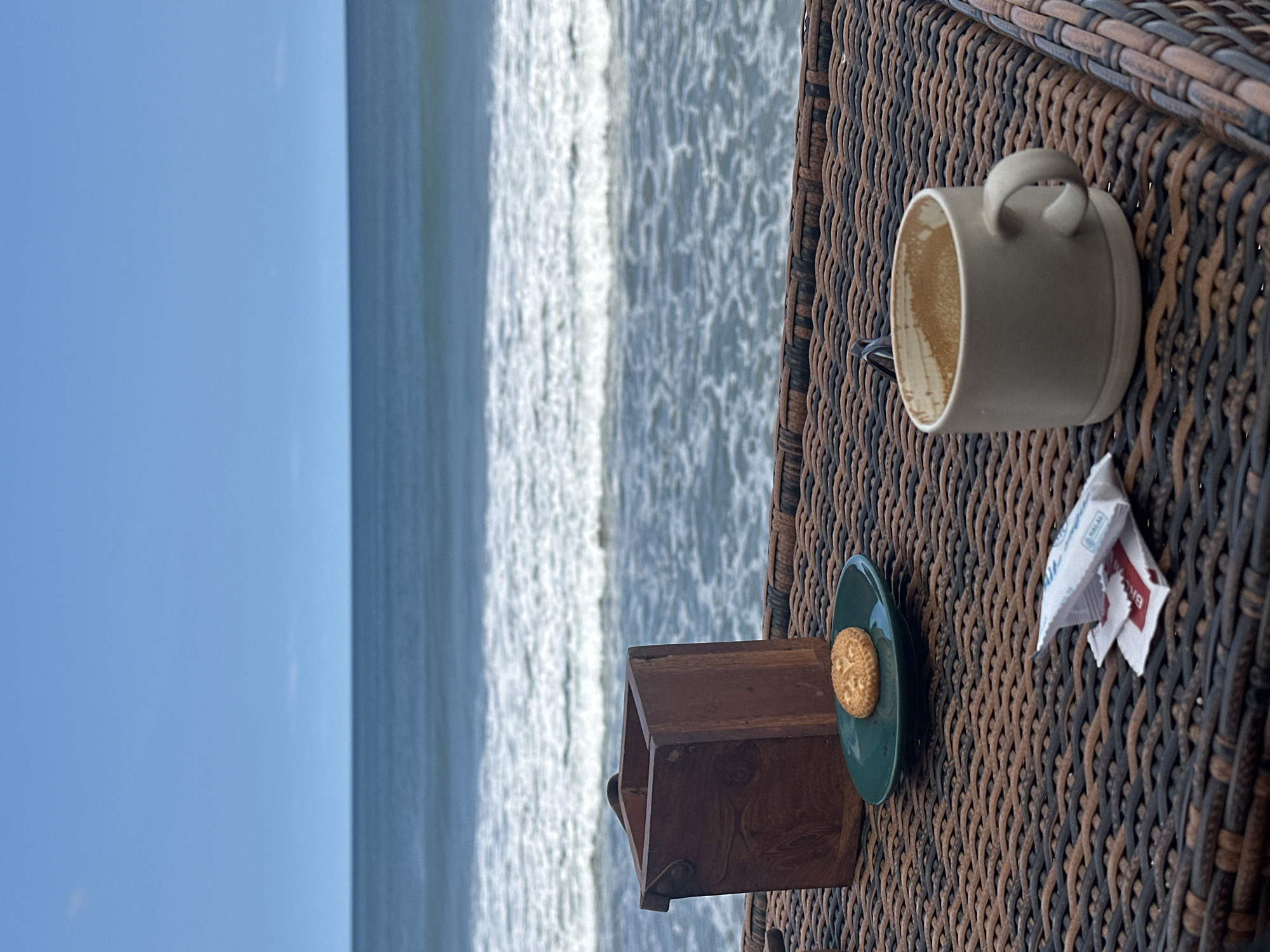 A table on a beach in North Kuta, Indonesia, holds a mostly empty coffee mug, a small green saucer with a biscuit, a small wooden box, and sugar packets. The ocean is in the background.