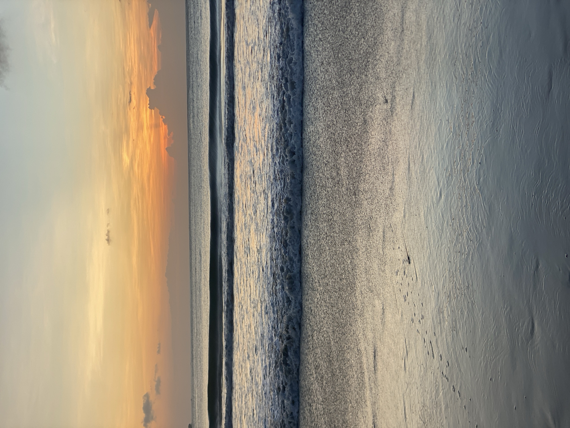 The image depicts a beach scene at sunset in Kuta Selatan, Indonesia. The sky is a mix of light grey and orange, with some cloud cover. The ocean has small waves and appears blue-grey in color. The sand is wet and grey, with patterns and lines imprinted on its surface. There are no visible people or objects on the beach.