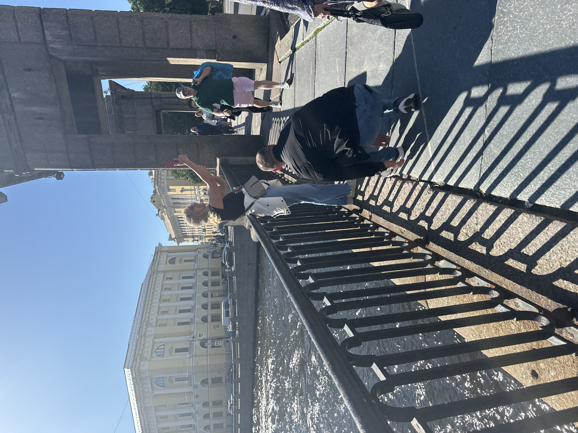 The image captures a scene on a sunny day in Saint Petersburg, Russia. In the foreground, a woman with blonde hair, wearing a black tank top and jeans, is captured in mid-motion, holding a pink phone and appearing to take a selfie or photograph. Beside her, a man in a black jacket and jeans is crouching, looking downwards. Behind them, a metal railing casts long shadows on the stone walkway, indicating the time of day is likely midday.
Further in the background, several other individuals are visible. One man, wearing a green shirt and pink shorts, stands with a blue bag slung over his shoulder. Other people are walking along the path, some in the distance. The setting appears to be a pedestrian area adjacent to a body of water, possibly a canal or river, reflecting the bright sunlight. Across the water, a grand, light-yellow building with classical architectural elements, characterized by columns and arched windows, is visible. The clear blue sky suggests fair weather. The overall atmosphere appears casual and relaxed, with people enjoying a day outdoors.