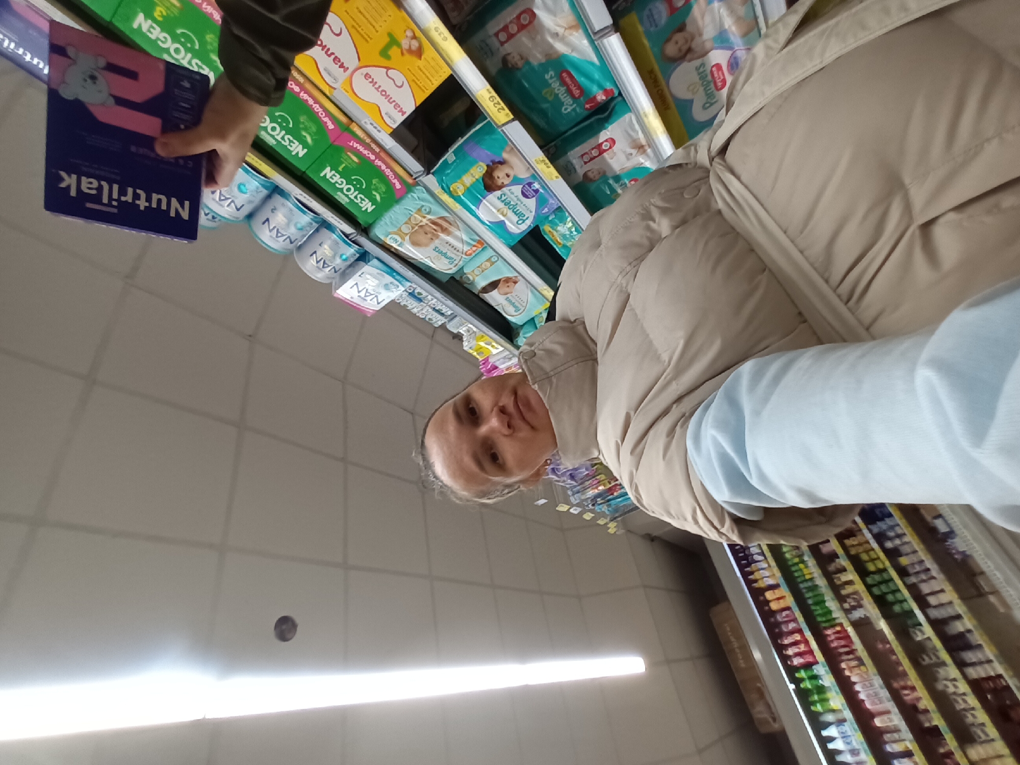 A woman in a beige jacket stands in a supermarket in Chaikovsky, Russia. She looks at the camera while holding a box of Nutrilak baby formula in her hand. Shelves of baby products, including Pampers diapers, Nestle Nan formula, and Nestogen formula, are visible. The shelves of groceries are also visible in the background.