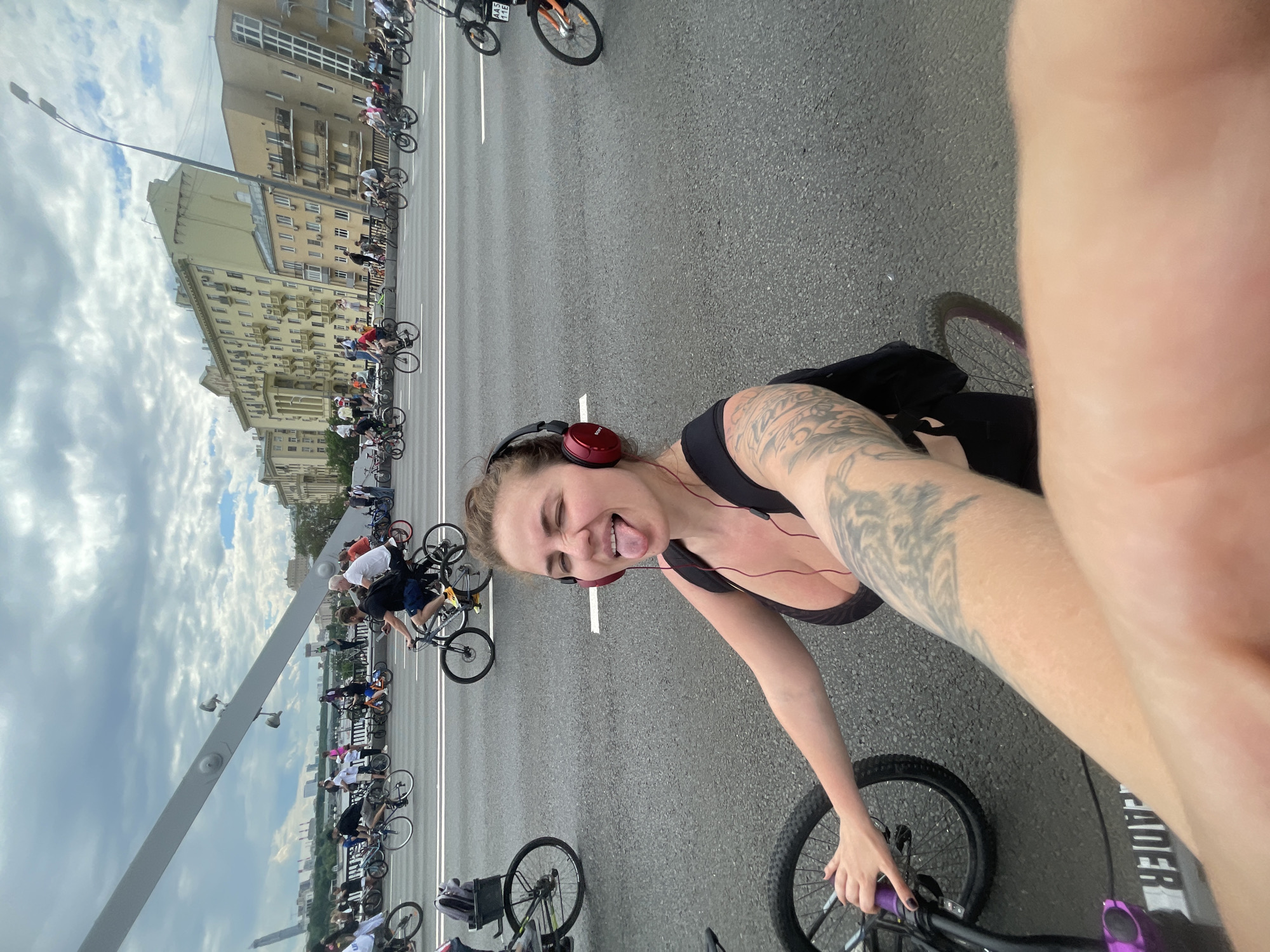 A slightly canted, medium shot depicts a woman on a bicycle amidst a cycling parade, purportedly in Moscow, Russia. She is wearing red headphones, a black tank top, and has tattoos on her left arm. Her tongue is sticking out, indicating a playful mood.
The background showcases a dense gathering of cyclists on a grey asphalt surface, presumed to be a road or bridge, marked with white lane dividers. In the distance, buildings are visible, consistent with urban architecture common in Moscow. The sky above is overcast. A street light is seen at the left of the frame.