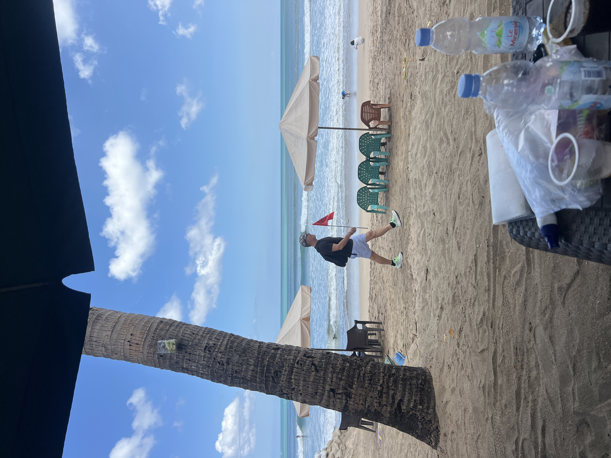 A man walks on a sandy beach in Kuta, Indonesia, with the ocean and waves in the background. He is wearing a black t-shirt, light blue shorts, a cap, and athletic shoes. He carries a black backpack and walks past green plastic chairs and a beige umbrella. To the left of the man, a large palm tree stands prominently. Further in the background, two people are near the water, and a red flag is planted in the sand. In the foreground, a table holds several plastic bottles of water and cups. The sky is blue with scattered white clouds.