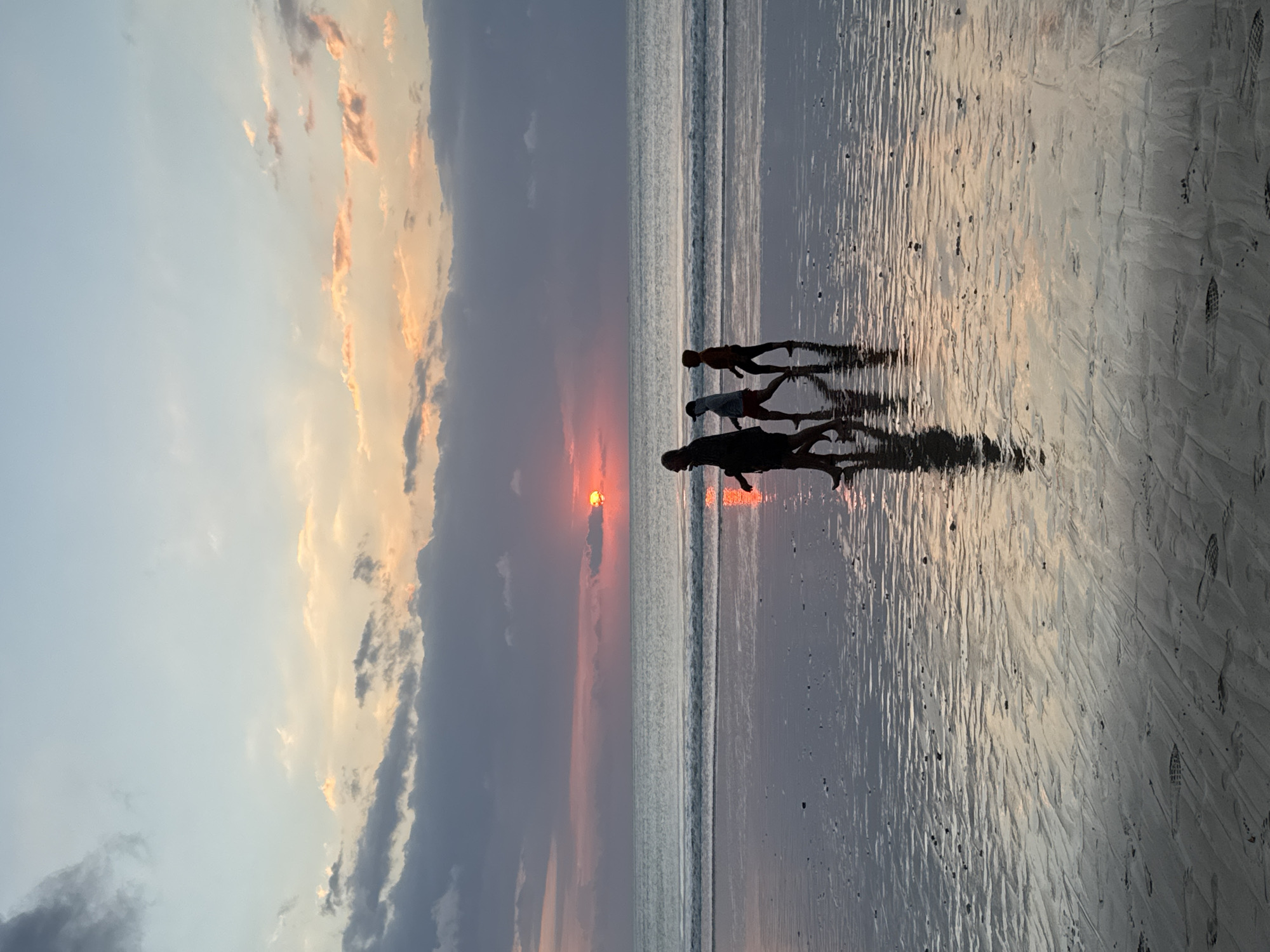 The image depicts a serene beach scene at sunset in Kuta Selatan, Indonesia. Three human figures are visible in silhouette, walking from left to right across the wet, reflective sand near the ocean's edge. Their dark forms are mirrored in the damp surface, creating elongated reflections. The sun, a vibrant orange-red disc, is low on the horizon towards the left, partially obscured by dark and brightly lit clouds that stretch across the sky.
The sky transitions from a soft blue-grey at the top to warmer hues of orange and pink near the sun, indicating either a sunrise or, more likely, a sunset. Gentle waves are visible in the ocean, forming horizontal lines of white foam across the water. The expansive wet sand reflects the colors of the sky and the bright sun, with scattered footprints visible further inland on the drier parts of the beach. The overall atmosphere is tranquil and picturesque, capturing a moment of peaceful contemplation by the sea. There is no visible text in the image.