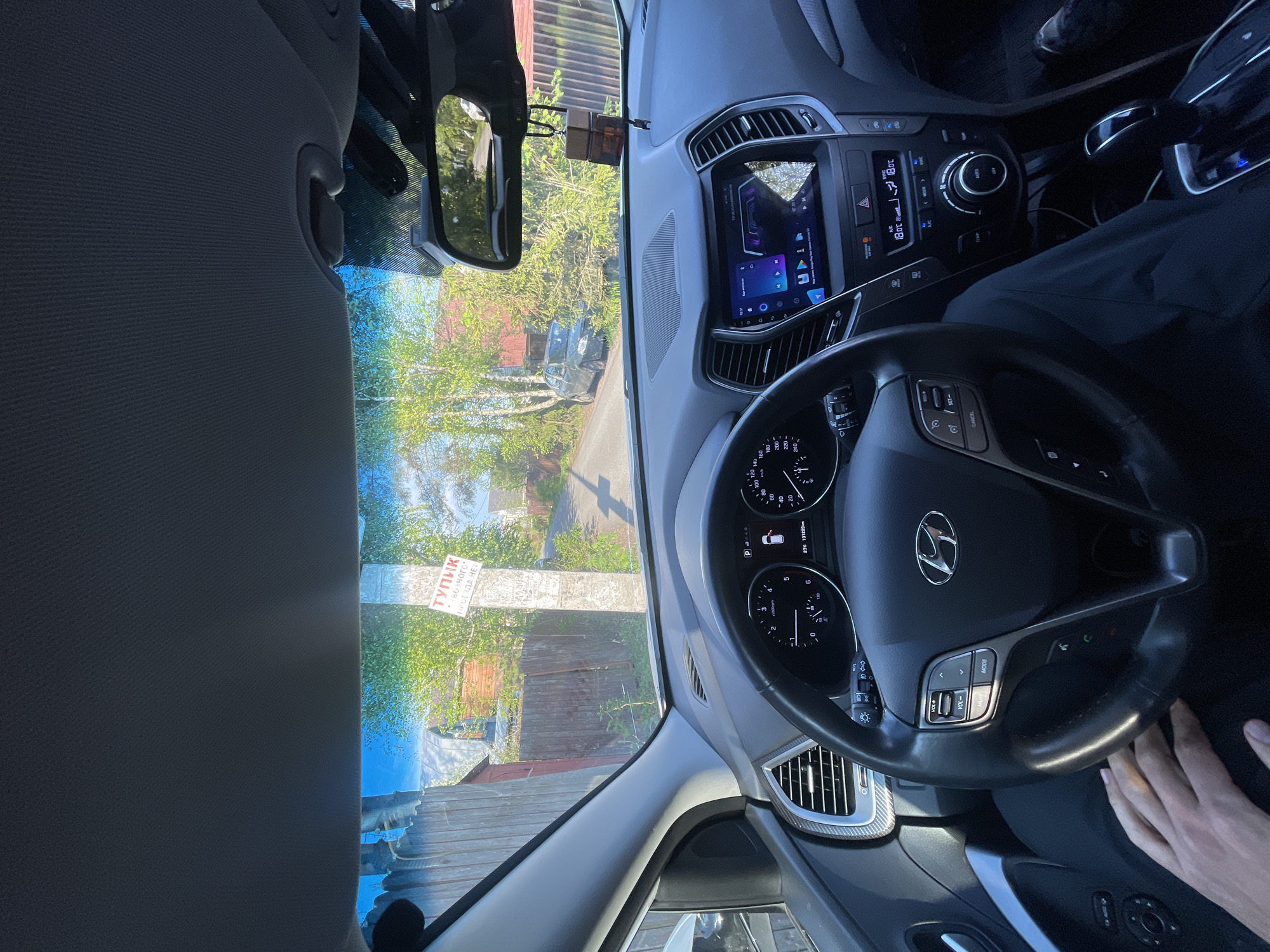 Here's a description of the image:
The image shows the interior of a car, specifically the driver's seat, with the dashboard, steering wheel, and part of the windshield visible. The car appears to be a Hyundai, based on the logo on the steering wheel. The dashboard features a digital display and various controls for the car's systems. A person is sitting in the driver's seat with their hand visible on the steering wheel.
Through the windshield, a street scene is visible. There is a sign written in Russian that says "Тупик", which translates to "Dead End", and below is written "ВОЗНОГО ПРОЕЗДА НЕТ" which translates to "NO THROUGH PASSAGE". Based on the Cyrillic script, the location is likely in Russia. The sign is attached to a post near a wooden fence. The surrounding area has trees with green leaves, suggesting it is springtime. This location can be specified as Sestroretsk, Russia, based on the signs and surrounding landscape.