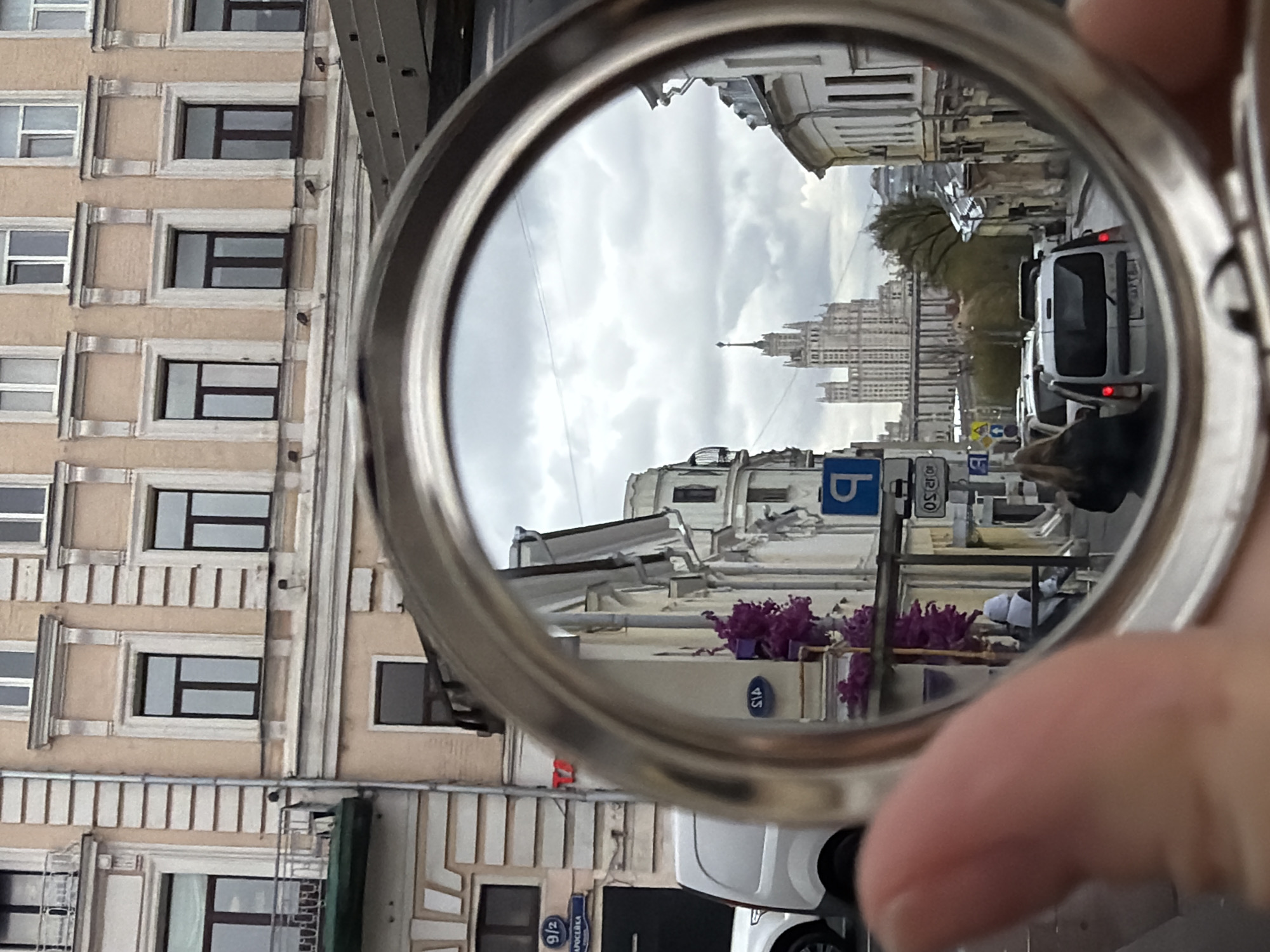 This photograph captures a Moscow street scene, partially viewed through a circular reflective object, likely a compact mirror or a magnifying glass. The image is oriented vertically, though the reflected scene suggests a horizontal street view.
Outside the reflective circle, a multi-story building facade occupies the left side, characterized by beige walls, rectangular windows with dark frames, and decorative pilasters or relief details. A hand is partially visible in the lower right, holding the metallic rim of the circular object.
Inside the reflection, a wide street stretches into the distance under an overcast, cloudy sky. Prominently in the mid-ground stands one of Moscow's iconic Stalinist skyscrapers, with its distinctive tiered architecture and spired top. Along the street are various buildings, some with traditional European architectural elements. Several white vans and cars are visible in what appears to be traffic, one showing its red taillights. Purple flowers in planters add a touch of color to the urban landscape.
Visible text and signs include a blue "P" parking sign, a sign with "8|12|50" (likely indicating parking regulations), and a blue oval street sign displaying "51A". Below the main reflective view, outside the circle, another blue sign with "9/2 СУРОВСКА" (Surovska, a street name) is partially seen, further confirming the Moscow location. The scene depicts a typical daytime urban environment with grey weather, observed indirectly through a reflective lens.