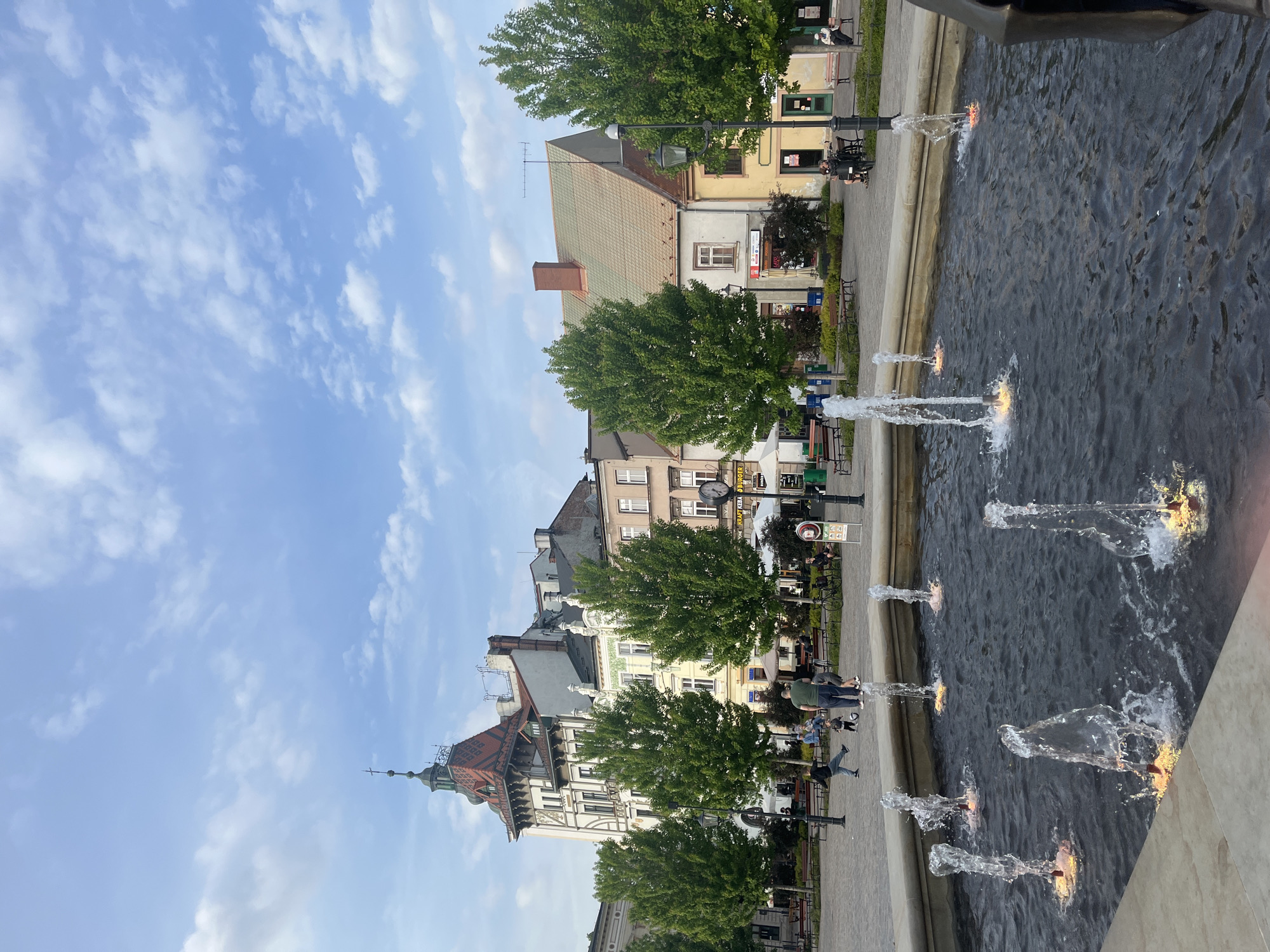 The image shows a cityscape with a water feature in the foreground. The camera angle is low, capturing a broad view of the buildings and sky.
The sky is light blue with scattered white clouds. Buildings of various heights and architectural styles line the street. Some have pitched roofs, while others are flat. The buildings are constructed of brick and concrete. Trees with lush green foliage are planted along the street, partially obscuring the buildings.
In the foreground, a water feature with multiple fountains is visible. Water is ejected upward from the fountains. People are walking and sitting on benches along the street. A man is walking along the edge of the water feature.