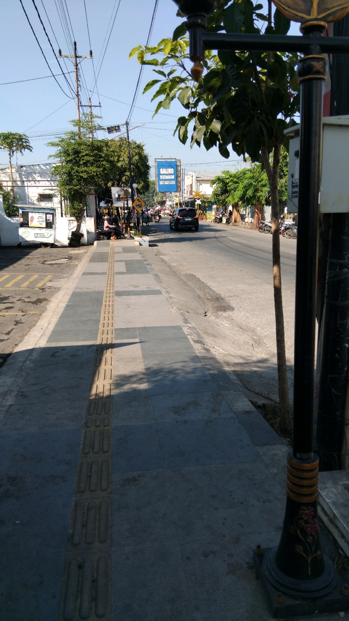 The image depicts a street scene in Batang, Indonesia, on a clear, sunny day. The foreground is dominated by a sidewalk with tactile paving strips, indicating a pedestrian walkway. Adjacent to the sidewalk is a paved road with moderate traffic, including several motorcycles and a car. Trees line the right side of the road, casting shadows on the pavement.
On the left side of the street, there are buildings and shops. One establishment features a sign with an ice cream advertisement. Further down the street, a large blue billboard is visible with Indonesian text. Several individuals are present, mostly on foot or on motorcycles, suggesting a bustling urban environment. A lamppost with decorative elements stands on the right side of the sidewalk in the foreground. The overall impression is that of a typical day in a commercial area of an Indonesian city.