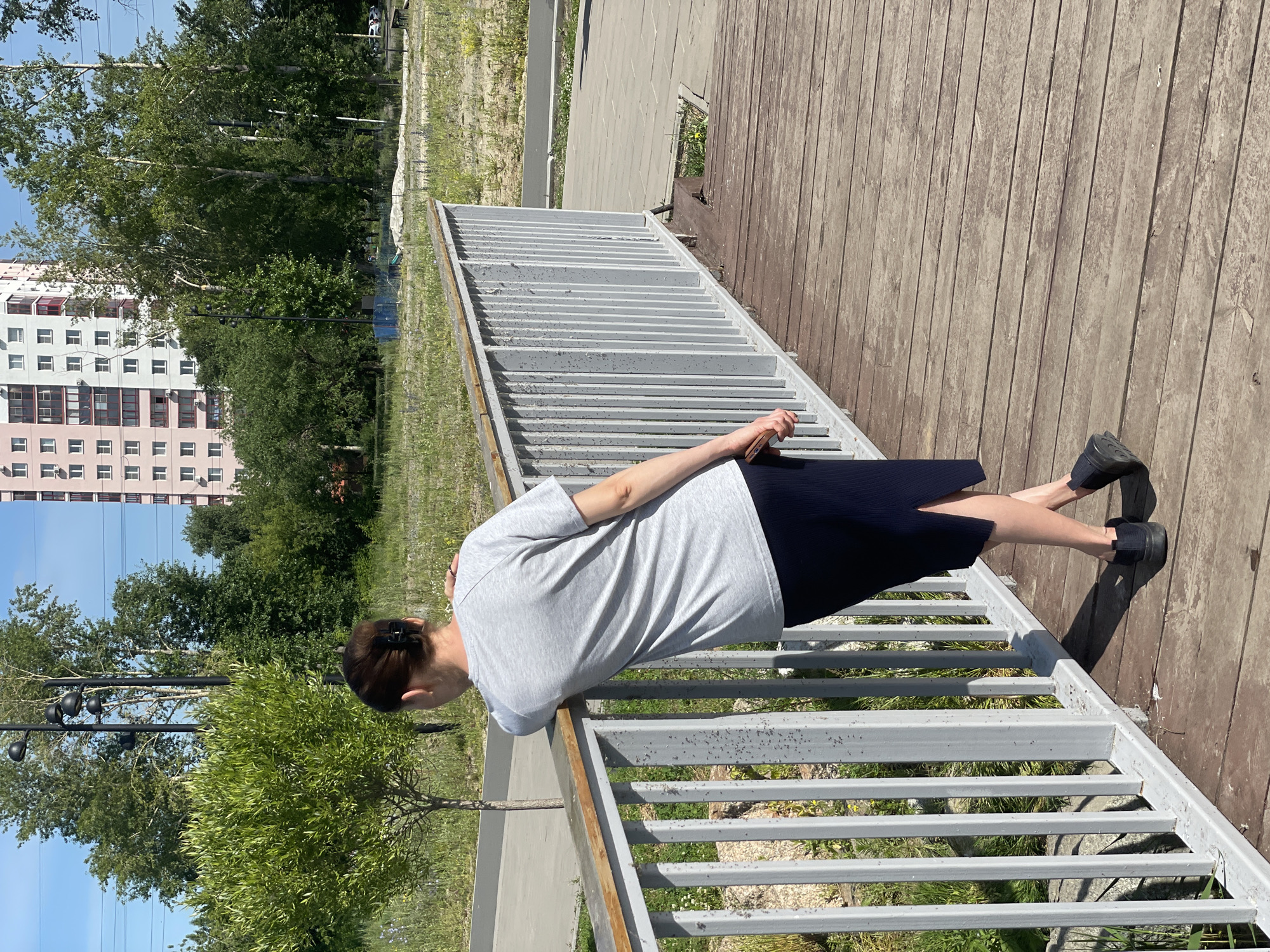 A woman stands on a wooden bridge, leaning on a white metal railing. She is viewed from behind, wearing a gray t-shirt and a dark blue skirt. Her hair is pulled back, and she is wearing black shoes. The bridge overlooks a grassy area with trees and a modern, multi-story building in the background. The sky is clear and blue.