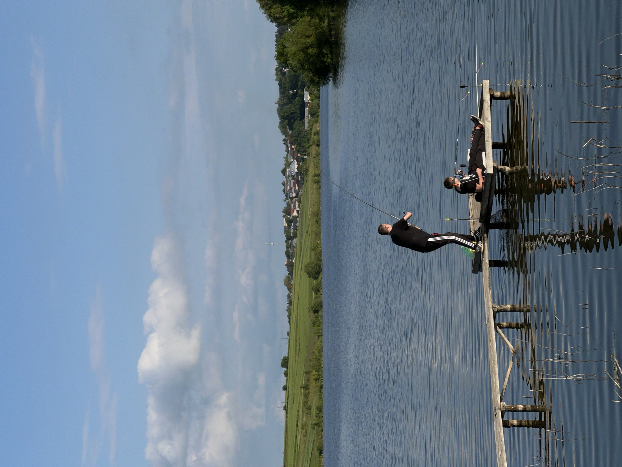 This image captures a serene daytime scene of two individuals fishing from a wooden pier on a calm body of water in Bazaryntsi, Ukraine.
In the foreground, a person dressed in dark clothing and white-striped athletic pants stands on the pier, actively holding a fishing rod extended over the water, appearing to be casting or reeling. Further along the pier, another person, also in dark attire, is seated or reclining, seemingly observing or waiting. Several fishing rods are visible on and around the pier, indicating a dedicated fishing activity.
The water is smooth with subtle ripples, reflecting the blue sky and providing a mirror-like surface. The wooden pier itself is rustic, constructed from planks and posts. Along the distant shoreline, a picturesque rural landscape unfolds, featuring expansive green fields and a scattering of houses that form a small village, partially obscured by trees. The horizon is low, allowing for a vast expanse of bright blue sky adorned with soft, white clouds.
The weather appears clear and sunny, suggesting a pleasant day for outdoor activity. The overall ambiance is tranquil and leisurely. No visible text is present in the image.
