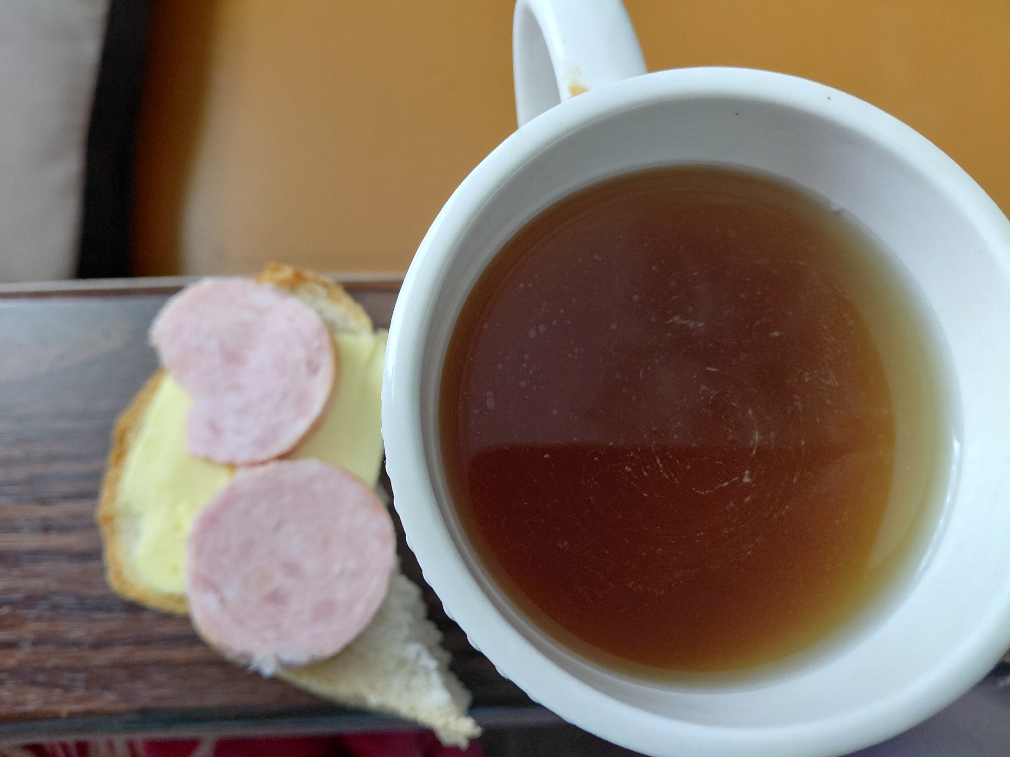 A close-up shot displays a white mug filled with a dark brown liquid, likely tea or coffee, positioned in the foreground. A slice of bread with butter and two circular slices of pink sausage, resembling ham, is visible in the background, resting on a dark wooden surface. The scene is set indoors, with an orange-toned surface occupying the right side of the frame. The image is taken from a high-angle perspective, emphasizing the contents of the mug. There is no indication of the city of Malinovka, Russia, or any specific location.