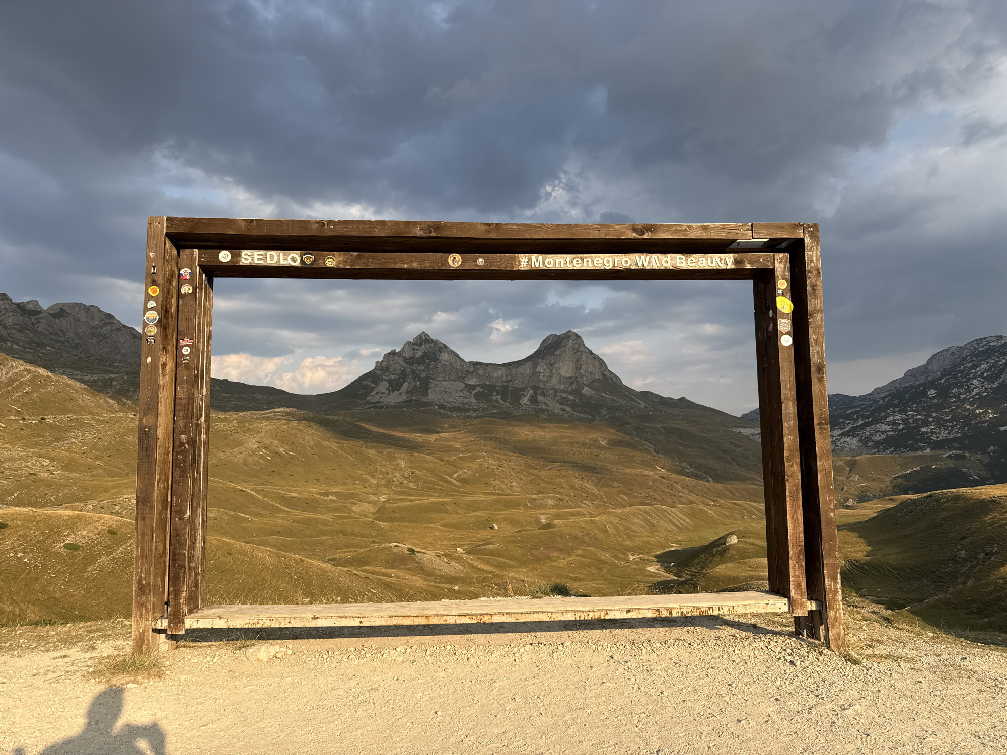 The image captures a view through a wooden frame, likely a scenic overlook or a photo booth, in Dubrovsko, Montenegro. The frame is adorned with several stickers and features the words "SEDLO" and "#MontenegroWildBeauty". Through the frame, a vast mountainous landscape unfolds under a dramatic sky with dark, heavy clouds interspersed with patches of lighter sky. The mountains in the distance are rugged and appear to be covered in sparse vegetation and rocky outcrops. The foreground consists of a sandy or gravelly path and the wooden structure itself, which has a bench attached to its lower section. The lighting suggests it is either early morning or late afternoon, with the sun casting long shadows, including the silhouette of a person holding a camera in the lower left corner. The overall scene conveys a sense of wild, untamed natural beauty.
