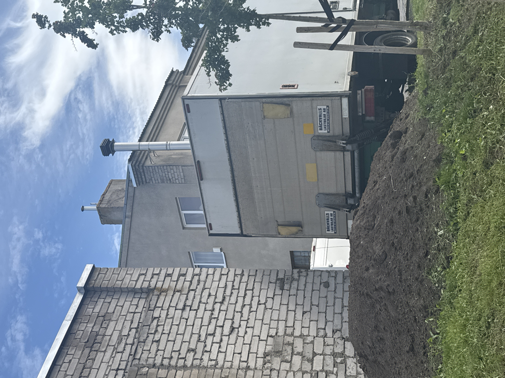 In Tallinn, Estonia, a scene unfolds with a pile of dark soil in the foreground, bordered by green grass. Behind the soil, a light-colored box truck with the branding "BÄCKVALLS LASTBILAR AB" is parked. The truck's rear lift gate is visible, along with a white van partially obscured behind it. To the left, a building constructed with white brick stands prominently. Further back, a grey building with windows and a chimney pipe rises against a sky filled with white, wispy clouds. A tree with green foliage is situated to the right of the truck.