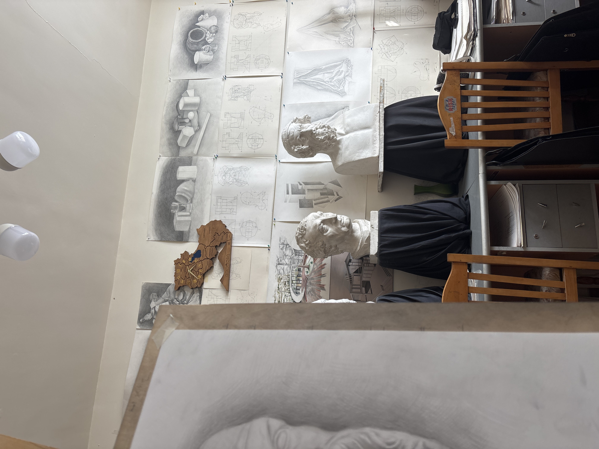 The image captures an art studio or classroom setting, viewed from a low angle looking towards a wall adorned with numerous drawings. The scene is dominated by a collection of pencil drawings pinned to a light-colored wall, showcasing various studies including still lifes of pottery and geometric objects, architectural or mechanical diagrams, drapery studies, and possibly figure drawings.
In the mid-ground, two classical-style white plaster busts of male figures with beards are prominently displayed. They appear to be resting on stands draped with black fabric, possibly serving as models for drawing exercises. One bust is positioned higher than the other.
To the right, a wooden chair with horizontal slats is visible, and on its back, a white sticker reads "I <3 My Tutor." Beyond the chairs, a grey metal shelving unit holds various papers and binders. A small green vase is also visible on one of the lower shelves.
In the lower left-mid section of the wall, a distinctive wooden, laser-cut map of Armenia is displayed, incorporating a clock face. Visible text on this map includes "ARTSAKH" and "ARMENIA." Two white LED light bulbs hang from the ceiling, illuminating the space. The ceiling/upper wall shows some faint cracks.
In the immediate foreground, a drawing board is partially visible, with a pencil drawing in progress, possibly depicting another bust or figure study. No people are visible, but the arrangement strongly suggests an active learning or practice environment for art. The setting implies a focus on observational drawing and technical studies within an educational context in Yerevan, Armenia.