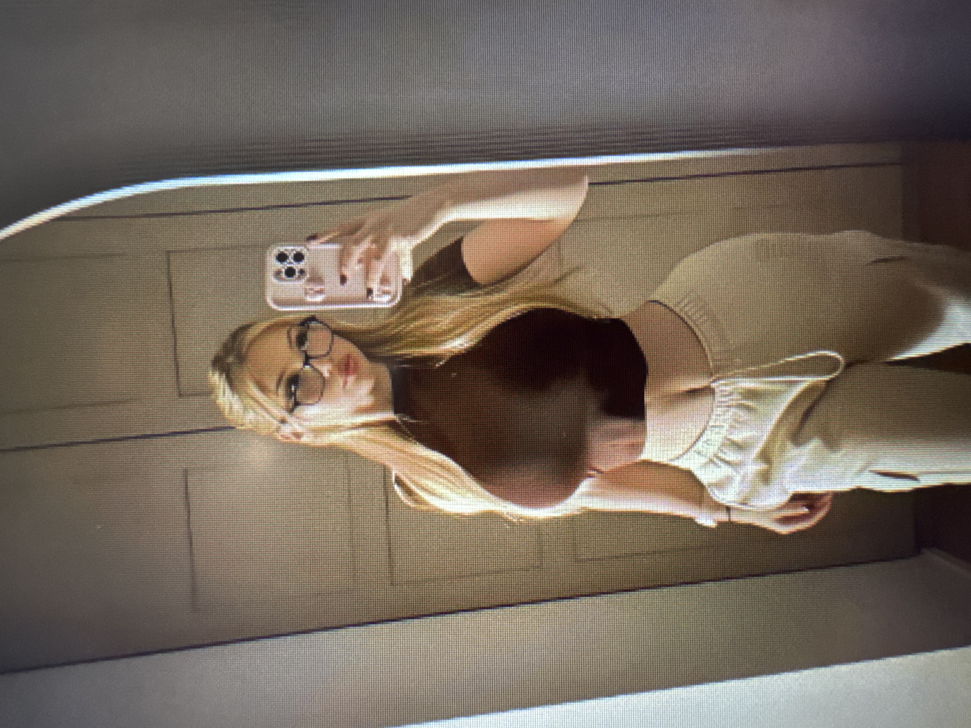 An image captures a young woman taking a mirror selfie. She is seen in a vertical orientation, holding a smartphone with her right hand to photograph her reflection. Her long blonde hair flows over her shoulders and down her back. She is wearing round-framed glasses and has a neutral or slightly serious expression. Her attire consists of a dark, form-fitting sleeveless top and light-colored, possibly beige or cream, loose-fitting pants, which appear to be sweatpants or joggers.
The smartphone she holds is light-colored, possibly pink or rose gold, and features a distinctive camera array with multiple lenses. A pop socket or grip is visible on the back of the phone. The woman's nails are painted a dark color.
The background visible in the mirror reflection is an indoor setting, likely a room with light-colored walls. There are visible vertical lines on the wall, suggesting paneling or doors, possibly a closet. The lighting is diffused, indicating an indoor environment. The overall image displays pixelation, suggesting it might be a photograph taken of a screen. There is no visible text in the image.