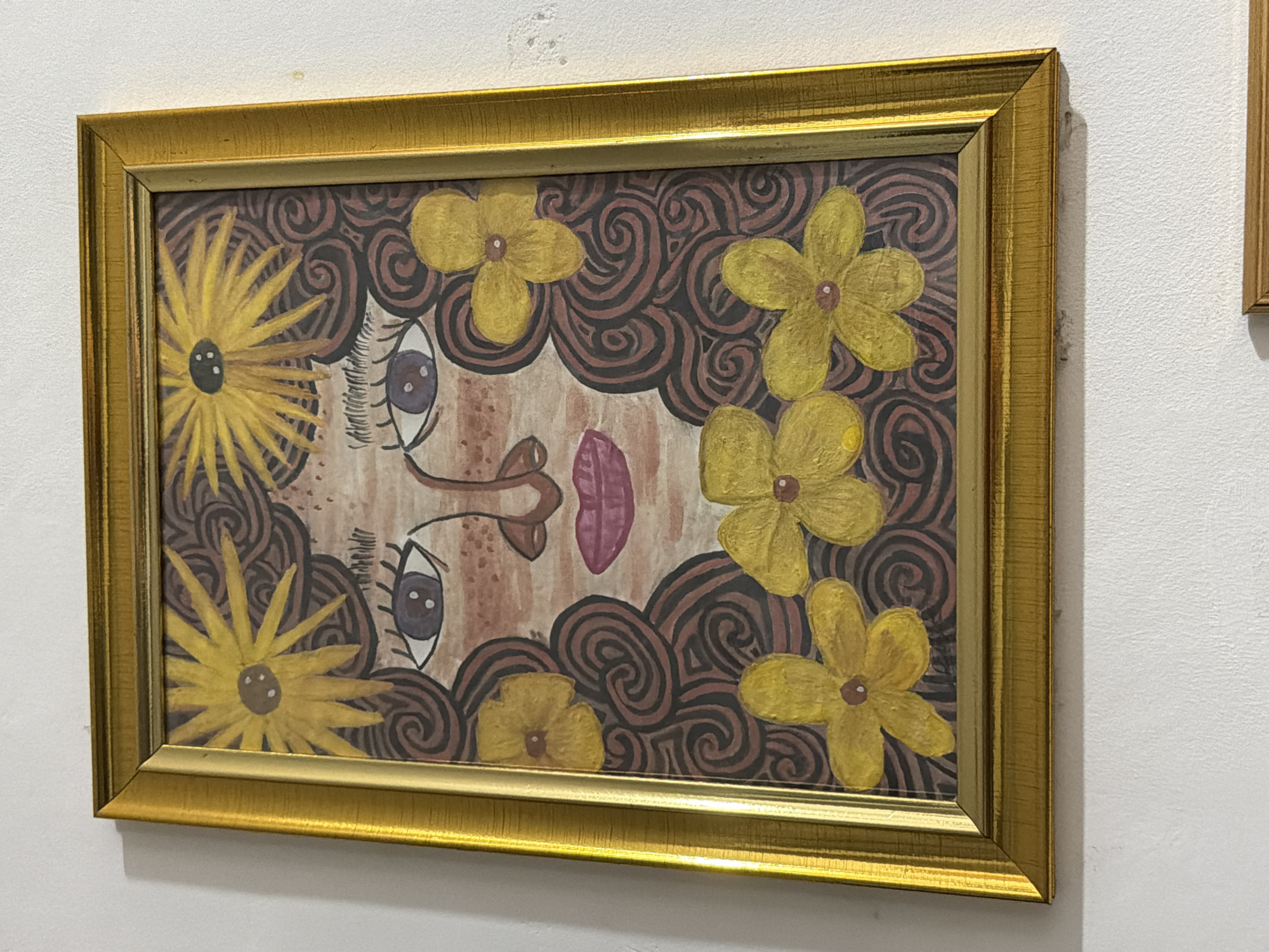 A framed painting is hanging on a white wall. The painting is a stylized portrait of a woman with curly brown hair, large purple eyes, and pink lips. Yellow flowers are incorporated into her hair and around her face. The frame is gold and has a textured finish. The wall appears to be painted white and shows some minor imperfections. There is also a sliver of another frame visible at the bottom of the image. No specific location in Davao City, Philippines can be determined from the image alone.