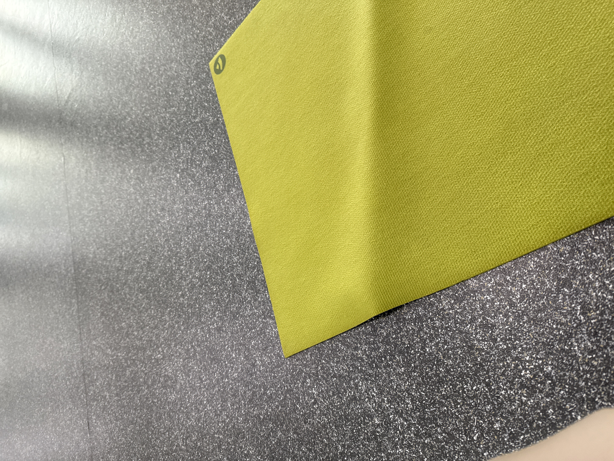 A close-up, slightly high-angle shot shows a section of a chartreuse-colored yoga mat lying on a mottled gray and silver floor. The yoga mat occupies the upper right portion of the frame, with a visible logo near its top left corner. The floor has a textured appearance, giving it a sparkling effect due to the silver particles. The scene is indoors, with the floor extending into the background. No people or other significant objects are present.