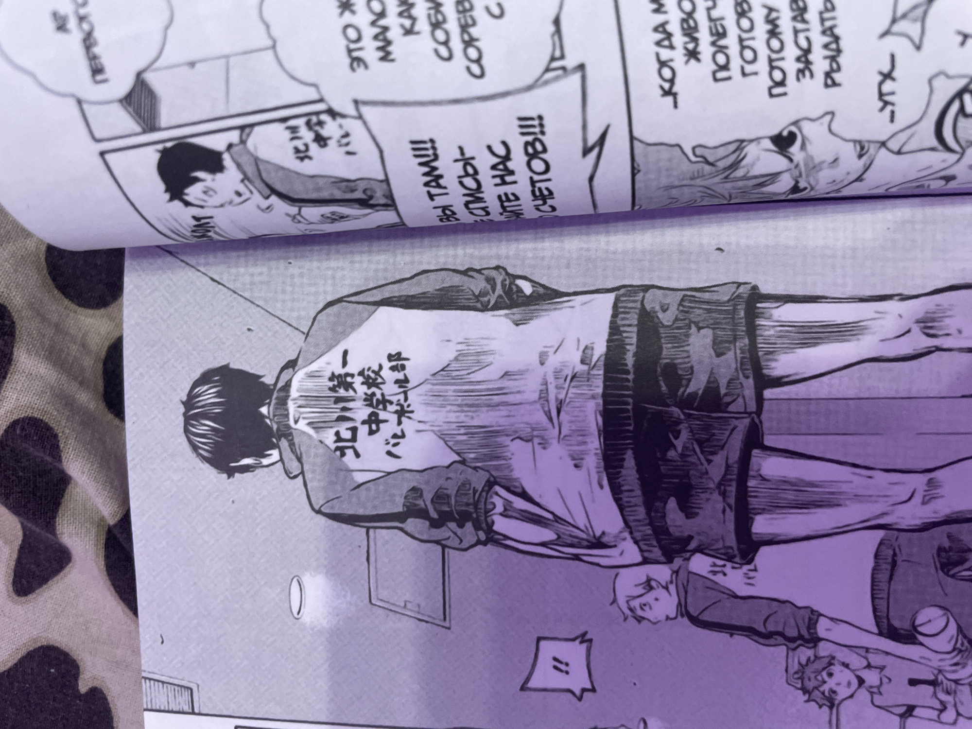 The image shows a close-up, slightly angled view of two manga comic book pages. The left page features a full-body illustration of a young man from behind, wearing a jacket with Japanese text on the back. He is facing away from the viewer, and a smaller figure is visible to his left, looking towards him. A speech bubble with "!!" is also present. The right page displays more manga panels with Japanese text and illustrations of two characters. The overall lighting has a purple hue, suggesting a filter or lighting effect applied to the photograph. The background of the image appears to be a patterned fabric, possibly a bedspread or blanket. There is no indication of the city of Irinovka, Russia, or any specific urban environment in this image.