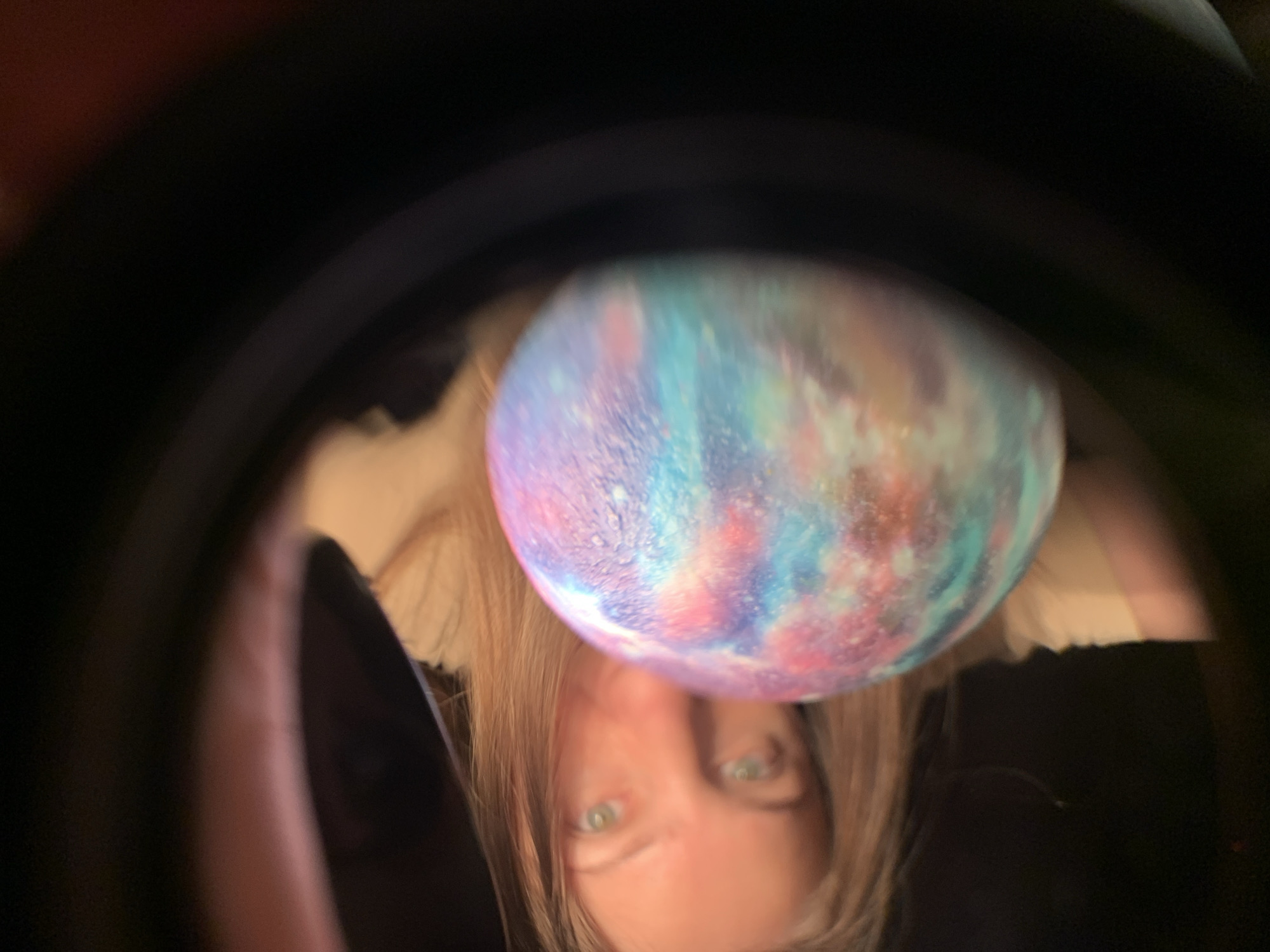 A person with blonde hair is looking down through a circular opening, possibly a lens or viewing device. The person's eyes and part of their face are visible through the opening. Above their face, within the same circular frame, is a colorful, spherical object, possibly a lamp or a decorative item, with a textured surface and shades of blue, purple, pink, and white. The surrounding area outside the circular frame is dark and out of focus. There is no information regarding the city of Chaikovsky, Russia.