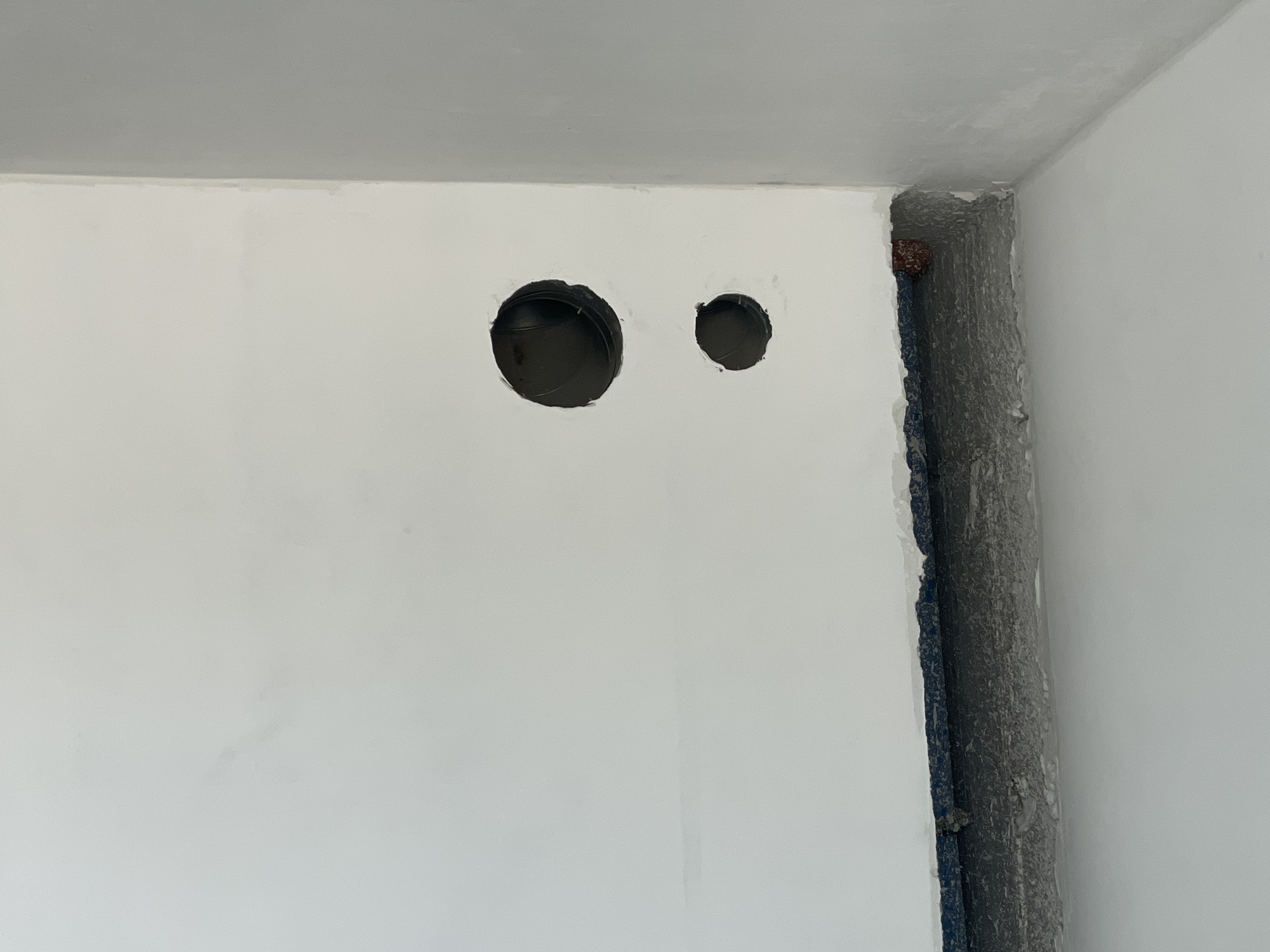 The image shows two black circular openings in a white wall. The openings appear to be roughly one foot apart. To the right of the openings, the wall is interrupted by a recessed area that reveals rough-textured gray material and a blue strip of some kind. The ceiling above the wall is a light gray color, and the walls appear to be freshly painted or plastered. The scene suggests an interior space under construction or renovation. The location is Unknown, Unknown.
