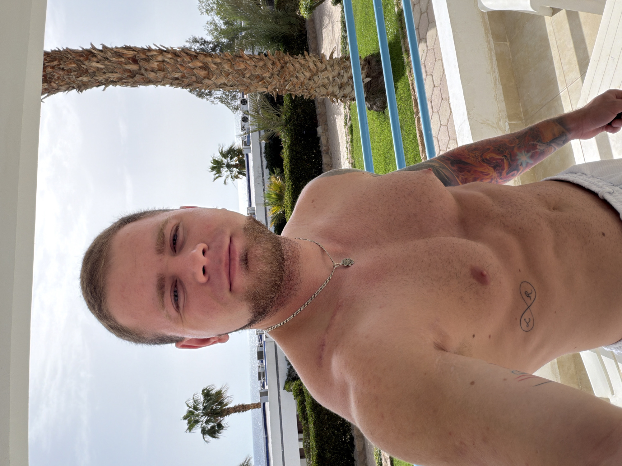 A man poses for a selfie outdoors in Sharm El Sheikh, Egypt. He has short hair, light skin, and a beard. He is shirtless, wearing a silver necklace with a pendant, and has tattoos on his arm and chest. The background shows palm trees, greenery, a building with a white facade, a light blue railing, and a partly cloudy sky.