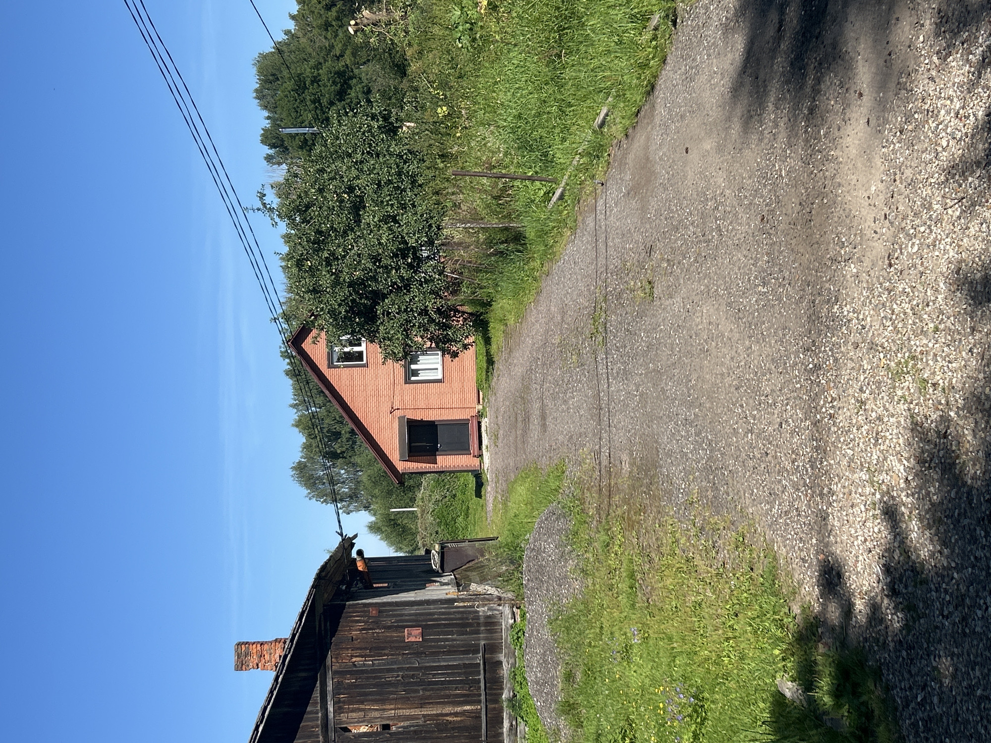 The image shows a rural scene in Kalitsino, Russia, on a clear, sunny day. In the foreground, a gravel driveway leads towards two buildings. To the left is a dark wooden barn with a brick chimney. To the right, a red brick house with white-trimmed windows and a dark door is visible. A mature tree with green leaves stands to the right of the house. The surrounding area is covered with green grass and some wild vegetation. Power lines are visible in the background against a clear blue sky. There are no people or animals present in the image.