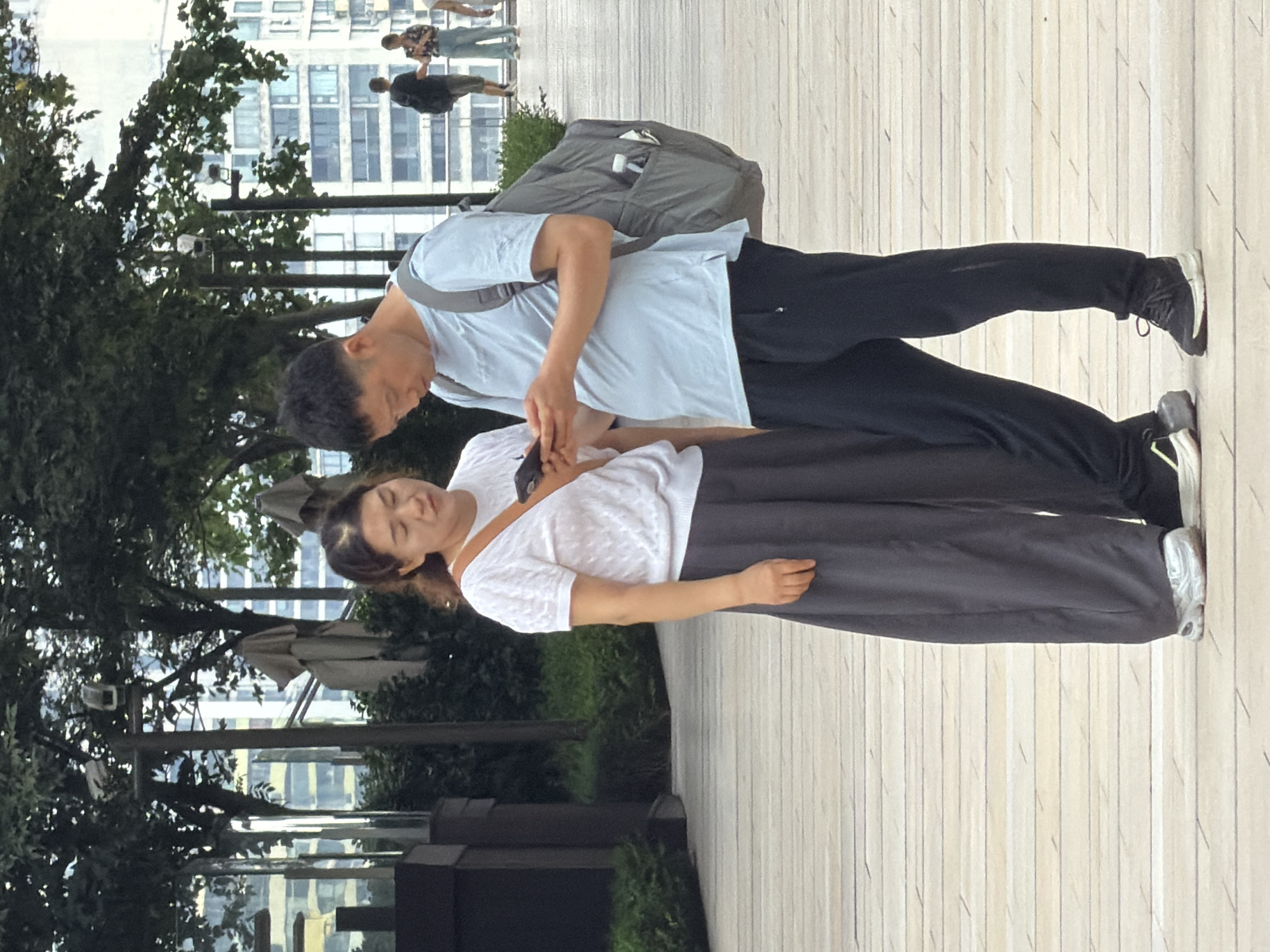Two individuals, a woman and a man, are standing on a wooden walkway outdoors. The woman, on the left, is wearing a white short-sleeved top with a textured pattern and dark grey wide-leg pants. She has a brown strap across her chest and is holding a dark-colored object, likely a phone, in her right hand. Her gaze is directed downwards. The man, on the right, is wearing a light blue t-shirt and black pants. He is carrying a grey backpack with a water bottle visible in a side pocket. He is holding a dark-colored object, also appearing to be a phone, in his hands and looking at it. Behind them, trees with dense foliage are visible, with a backdrop of modern buildings with many windows. The lighting suggests it is daytime. The environment appears to be a public urban space in Yuzhong District, China.