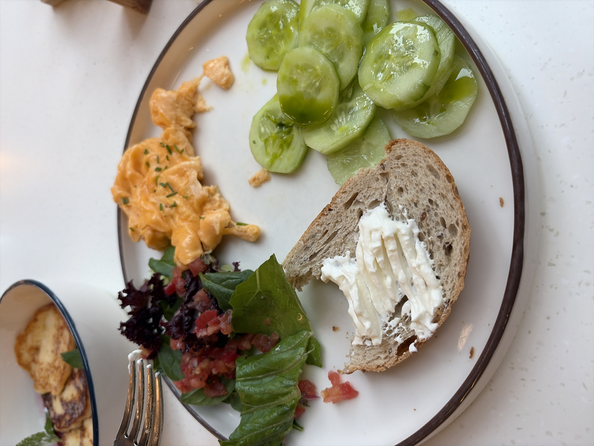 An overhead shot shows a white plate on a light-colored, speckled countertop. The plate contains a breakfast spread of scrambled eggs, a salad with tomatoes and greens, sliced cucumbers dressed with oil, and a slice of bread topped with cream cheese. A fork rests on the edge of the plate, touching the salad. In the upper left corner, a white bowl holds pieces of fried cheese and mint. The scene is lit from above, casting soft shadows. There is no indication of the specific location in Moscow, Russia.
