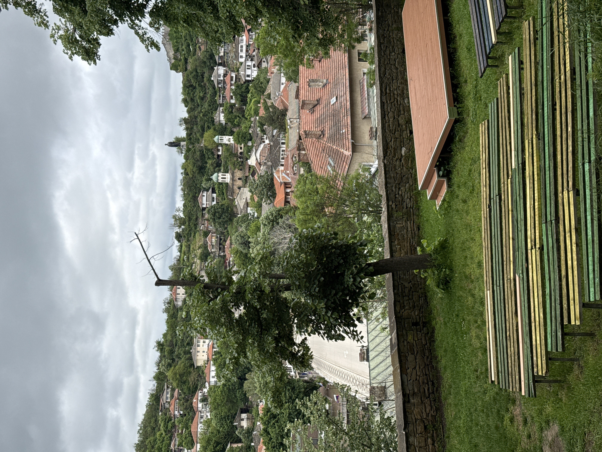 The photograph shows a hillside cityscape in Lovech, Bulgaria, under an overcast sky. Buildings with predominantly red-tiled roofs are densely packed and ascend the hill. The perspective is from an elevated position. To the right of the frame are wooden structures placed on a grassy area: two benches, one painted brown and another with a series of slats painted with green, yellow, and natural wood. A stone retaining wall is positioned alongside the benches and a tree. In the foreground, trees and foliage partially obscure the view of the cityscape.