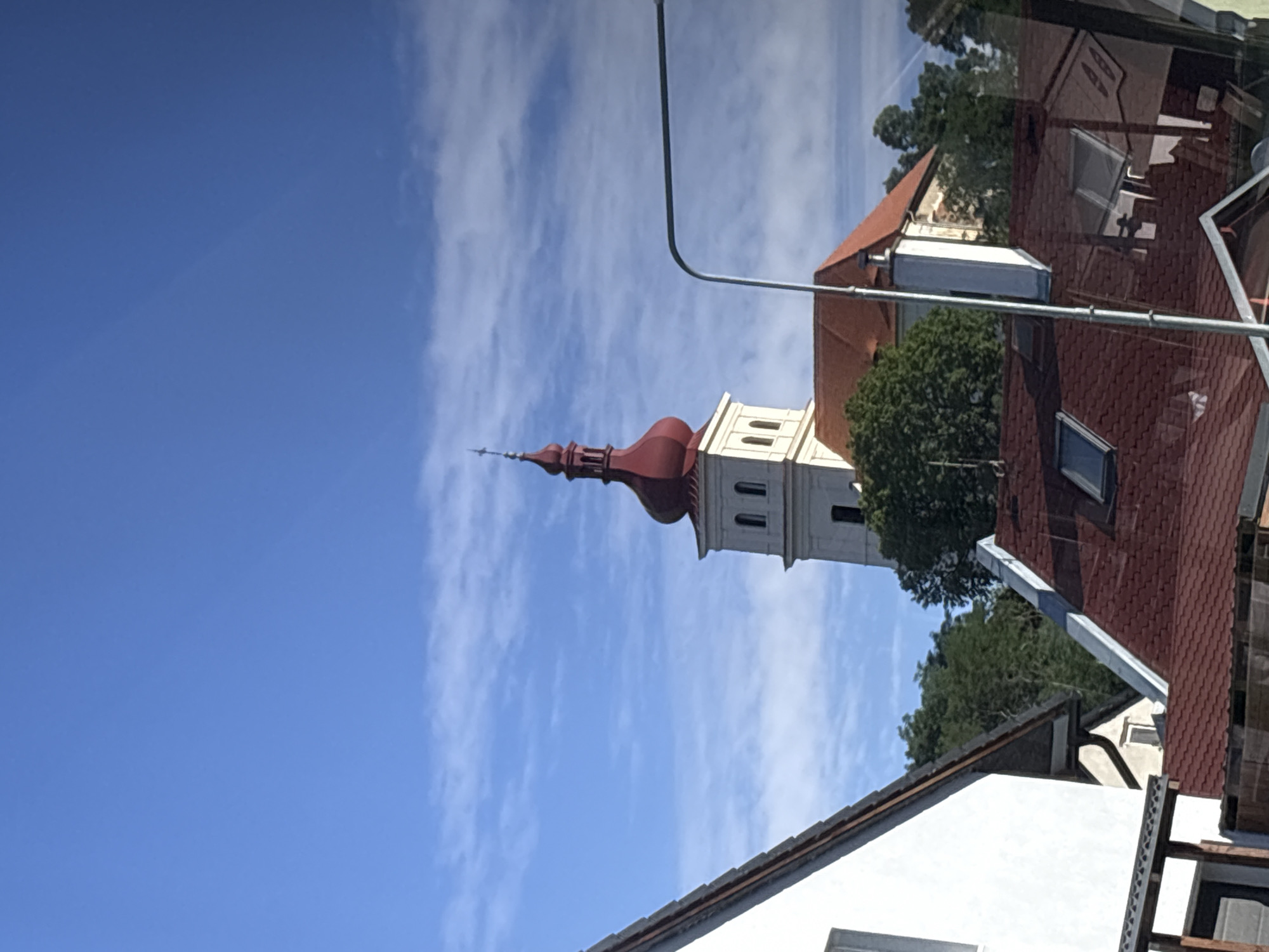A white church tower with a red onion dome and cross stands against a blue sky with scattered clouds. The tower has arched windows and a square base. In the foreground, several red-tiled roofs of residential buildings are visible, along with trees and a street lamp. The perspective suggests the photo was taken from a slightly elevated position, possibly from another building.