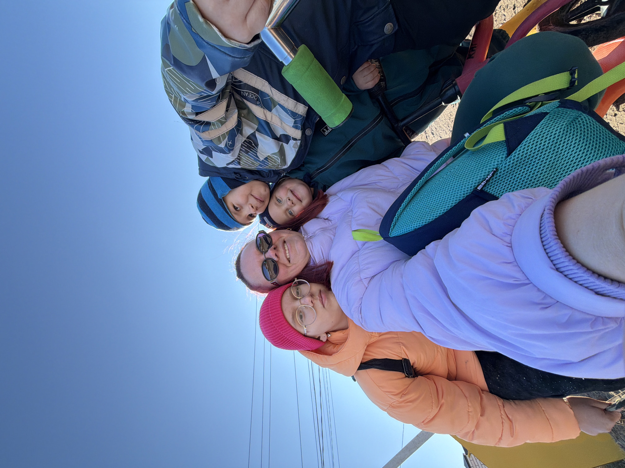 A group of five people are posing closely together for a photograph in the city of Chaikovsky, Russia. The group is oriented such that the sky is visible in the background. Two women are visible in the lower portion of the frame. One woman is wearing a pink beanie and a orange puffer coat and glasses. The other woman is wearing sunglasses and a lilac-colored puffer coat. Above the women are two small children, who are smiling. Above the two children is another figure wearing a patterned blue and green jacket and a striped blue beanie. Other accessories include backpacks and a green water bottle. Part of a vehicle with brightly-colored frame is visible in the upper right of the frame. Thin, grey wires intersect the sky above a pale yellow, semi-visible building.