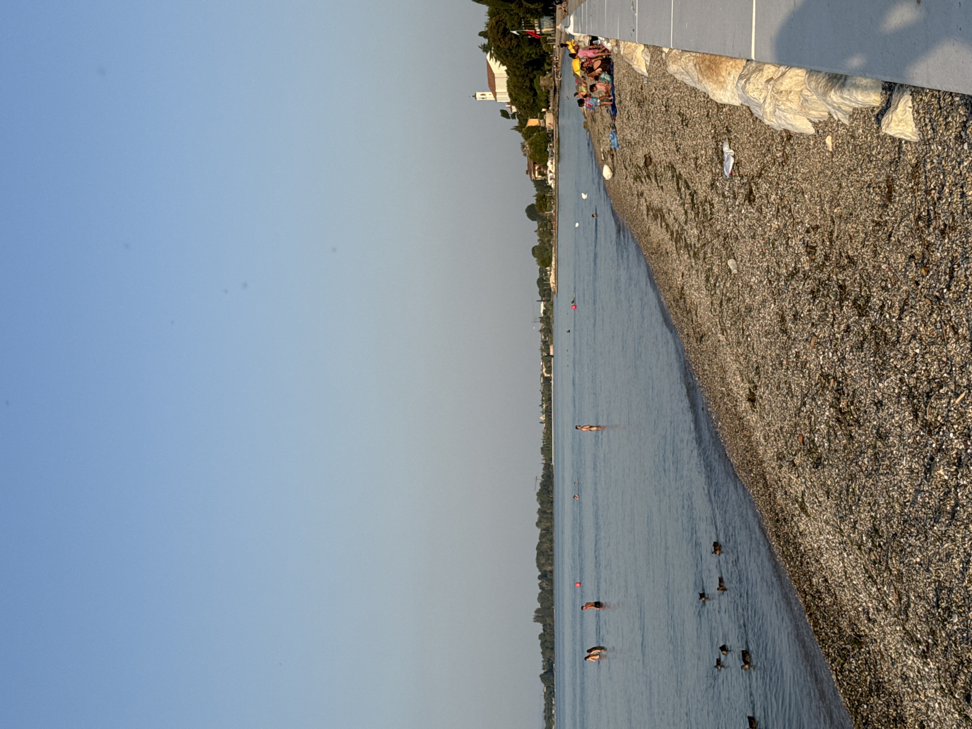 This media file captures a lively scene at a lakeside beach in Desenzano del Garda, Italy. The image is oriented vertically, with the sky occupying the left side and the beach and water on the right.
The subjects are a mix of people and waterfowl enjoying the day. Several individuals are visible on the gravelly beach, some sitting, others standing. A group of people is gathered near the water's edge, appearing to relax or prepare to enter the lake. Further out in the water, several people are swimming or wading. A flock of ducks is also present, swimming in the calm lake water.
The setting is clearly a lakeside beach with a paved walkway or promenade running along the top of the gravel beach. Behind the beach, a treeline is visible, with buildings, including a church with a steeple, suggesting a populated area. The time of day appears to be late afternoon or early evening, with the sun casting soft light and creating gentle shadows. The weather seems pleasant, with a clear sky and calm water.
Notable details include the texture of the gravel beach, the clear blue of the lake, and the bright clothing of some of the people. The presence of a church indicates the location is likely a town or city situated on the lake. The overall mood conveyed is one of leisure and enjoyment of a summer day. There is no visible text in the image.