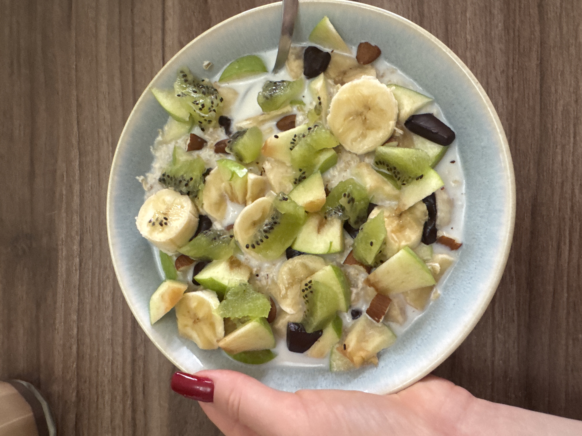 A light blue ceramic bowl, held by a human left hand, rests on a light brown wooden surface with horizontal grain. The bowl contains a breakfast meal comprised of a white liquid base, visible oats or cereal, sliced bananas, diced green apples, diced kiwi fruit, scattered dark brown chunks (likely chocolate), and lighter brown elongated pieces (likely almonds). A silver spoon is partially submerged within the contents on the right side of the bowl. The hand features a dark red painted thumbnail. In the upper left background, a blurred light brown object with a dark base is partially visible. The specific location within a city of Unknown, Unknown, is unidentifiable; the scene depicts an indoor setting.