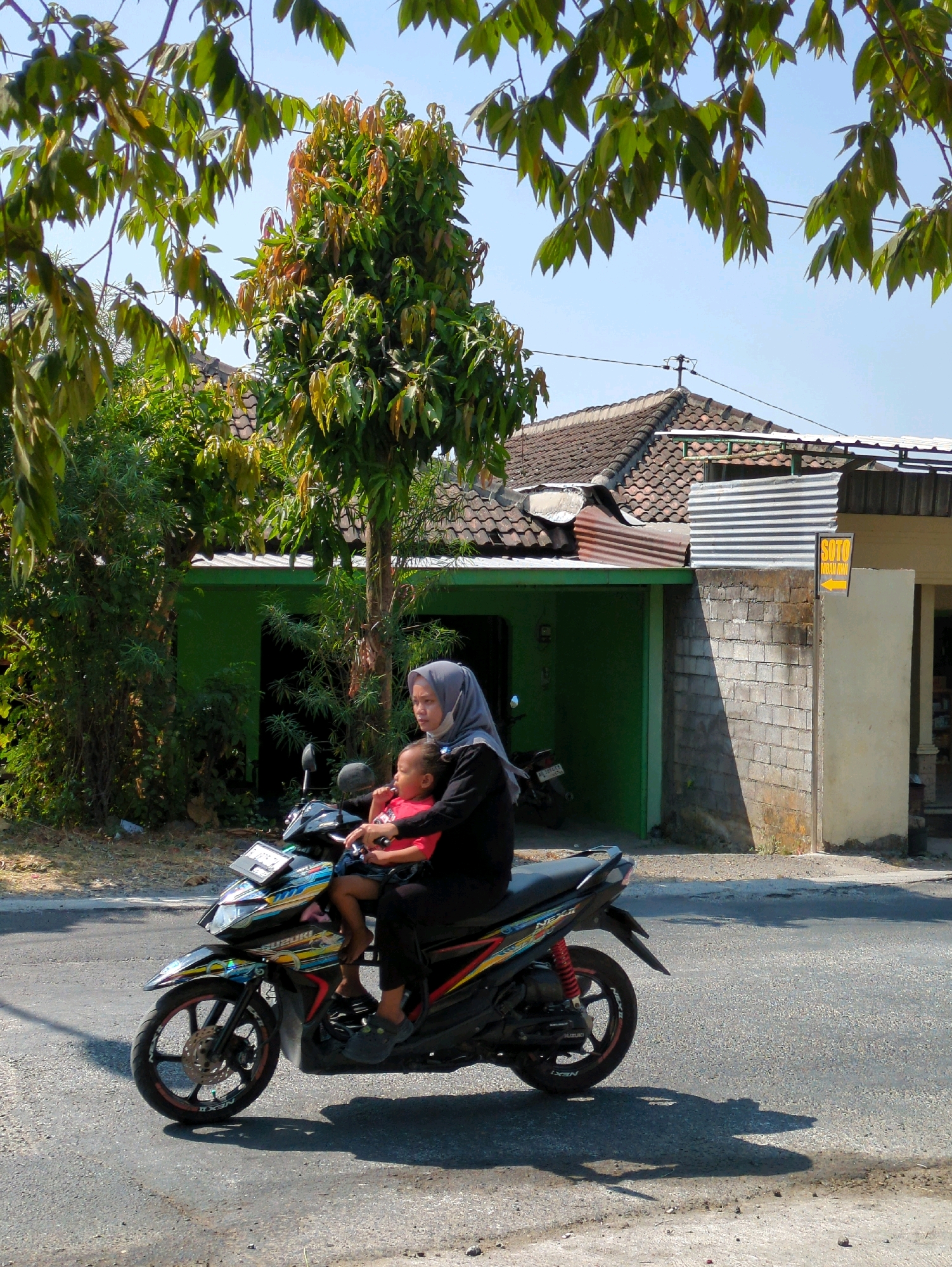 In Sindon, Indonesia, a woman wearing a grey hijab and black clothing rides a colorful motorcycle with a young child in a red shirt seated in front of her. The woman is looking to her right, while the child is looking forward. The motorcycle has a license plate that reads "AD 2442 CP". They are on an asphalt road with a shadow cast by the motorcycle and its occupants. To the left of the road, there is a lush green area with trees and foliage, and a vibrant green building with an open doorway. Behind the building, a house with a tiled roof is visible, with a sign that says "SOTO" with an arrow pointing to the right. The sky is a clear blue.